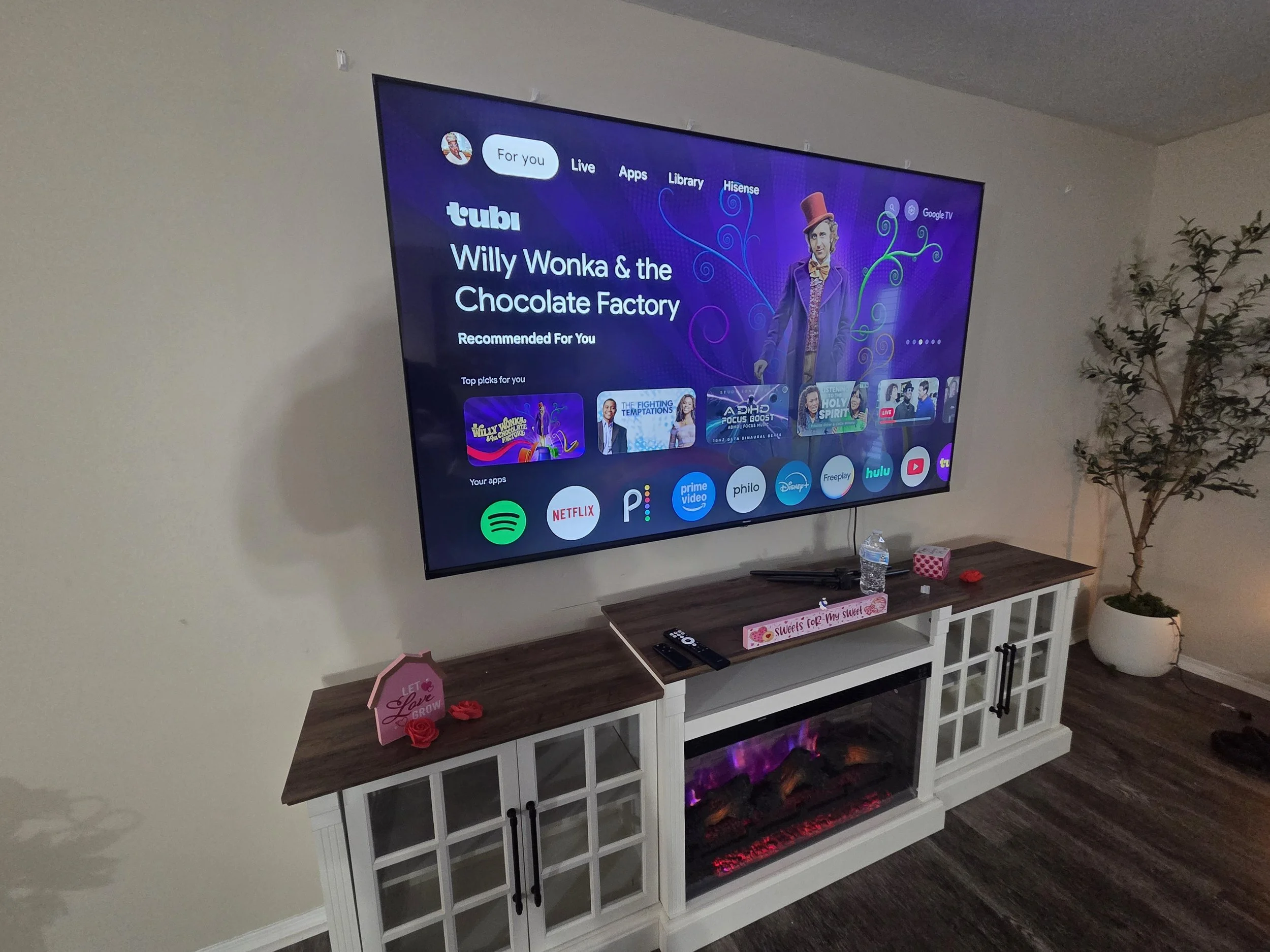 Living room entertainment area with a large flat-screen TV mounted on the wall, displaying the Netflix interface featuring Willy Wonka & the Chocolate Factory. Below the TV is a white entertainment console with two glass-paneled cabinets and an elect