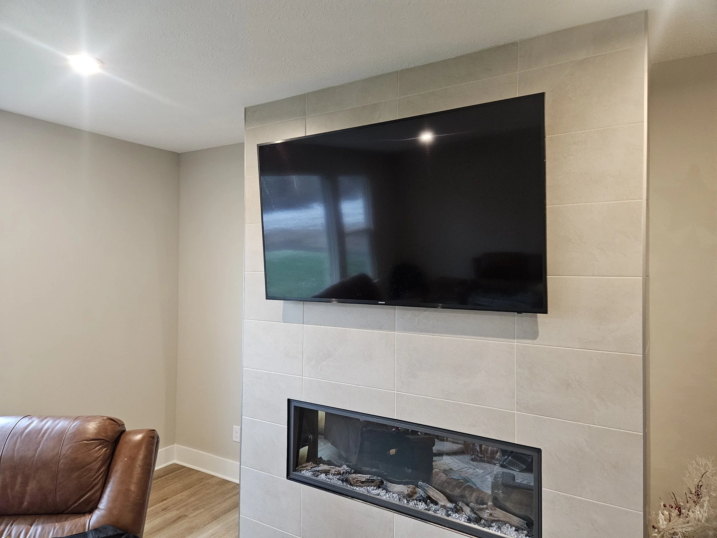 Living room with wall-mounted flat-screen TV above a modern fireplace with glass enclosure, beige tiled wall, and a brown leather chair in the foreground.