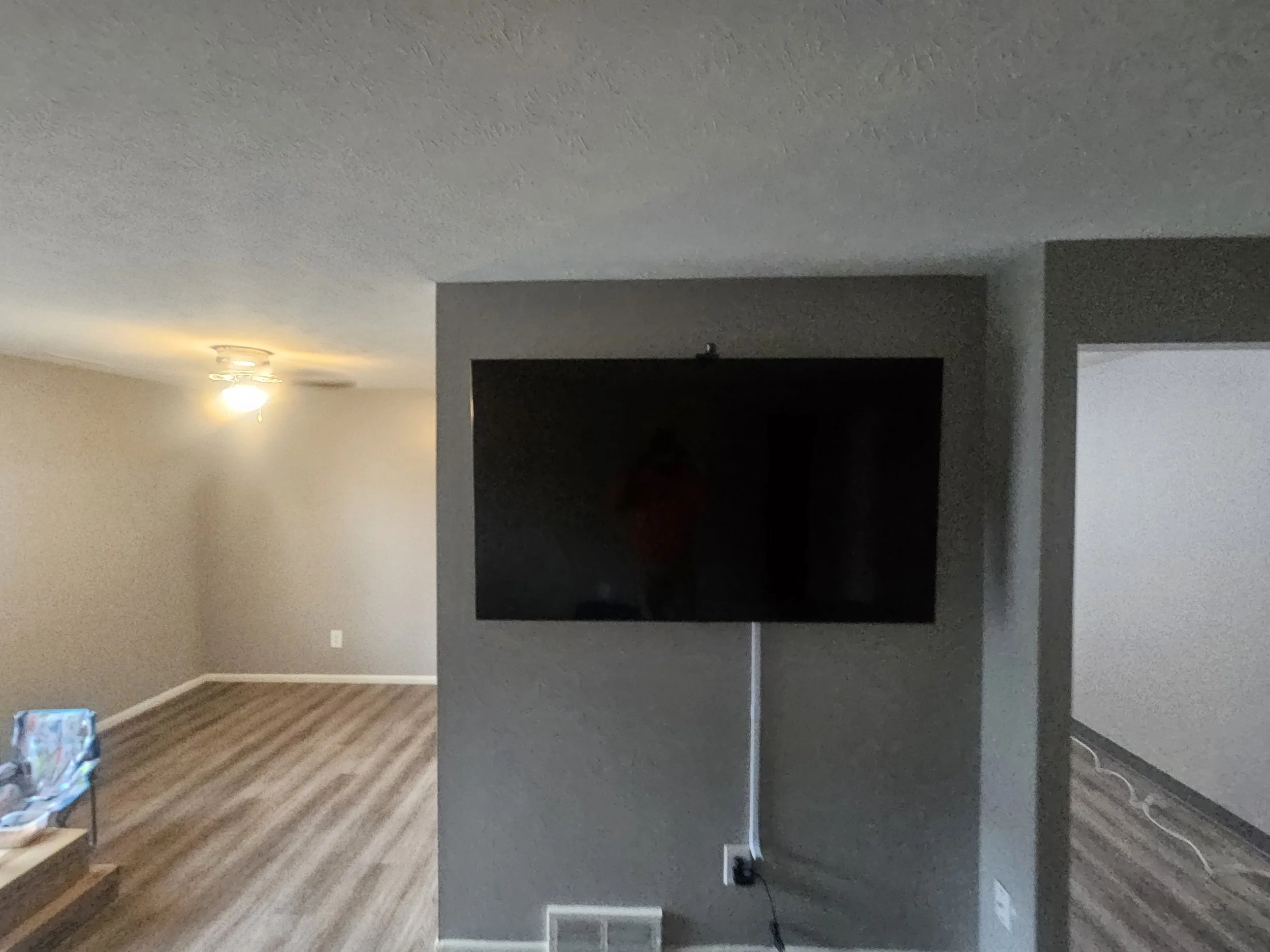 Living room with a wall-mounted flat-screen TV, hardwood flooring, a ceiling light, and a small side table with a colorful bag or object on it.