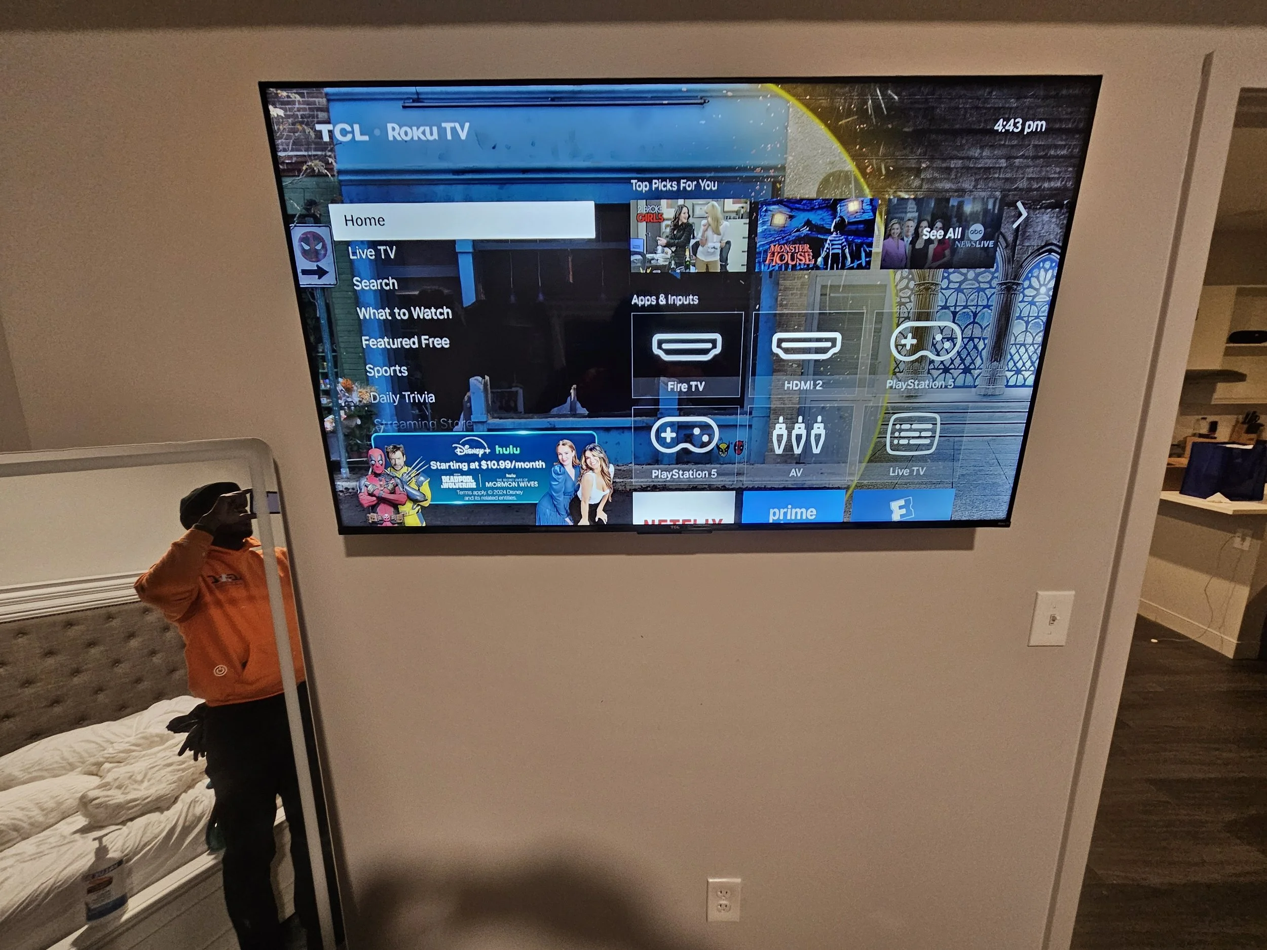 A television screen displaying a TV menu with options like Home, Live TV, Search, What to Watch, and more. The background shows a scene with blue structures and some promotional content at the bottom. A person in an orange hoodie is taking a photo of