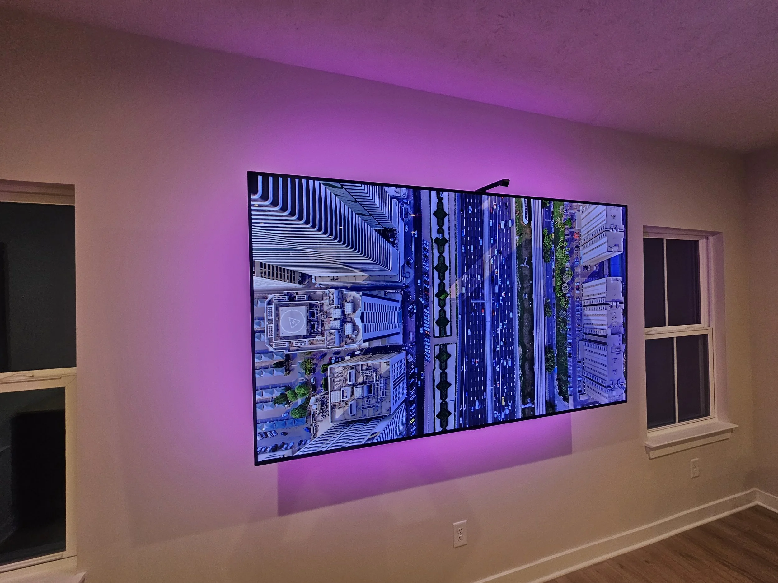 A flat-screen television mounted on a wall in a room, displaying a cityscape with tall buildings and roads, with purple ambient lighting
