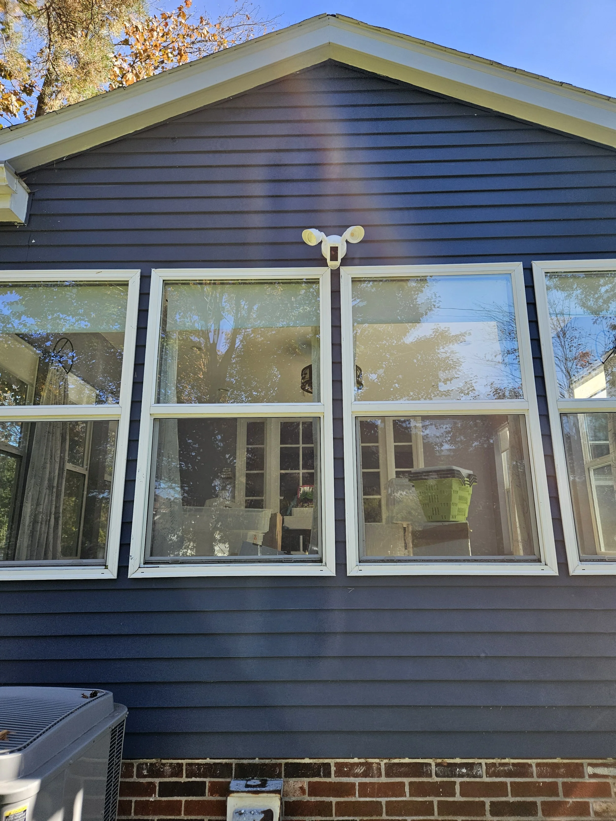 The back of a blue house with a gable roof, three sets of windows, and an exterior security camera mounted above the windows.