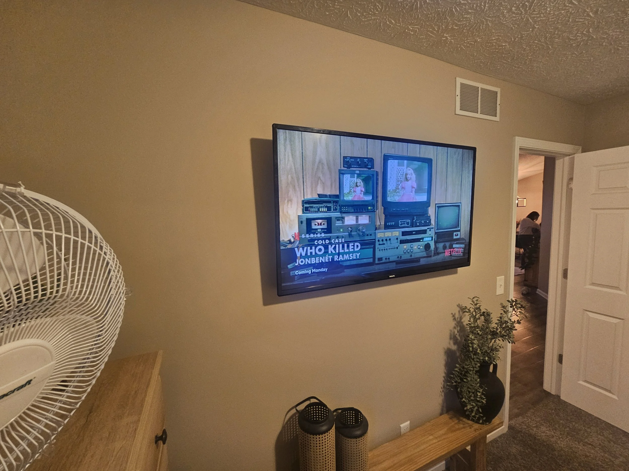 Living room wall with mounted television displaying a news story, a wooden bench with plant and decorative items, a large white fan on a wooden surface, and an open door revealing a person seated in a room in the background.