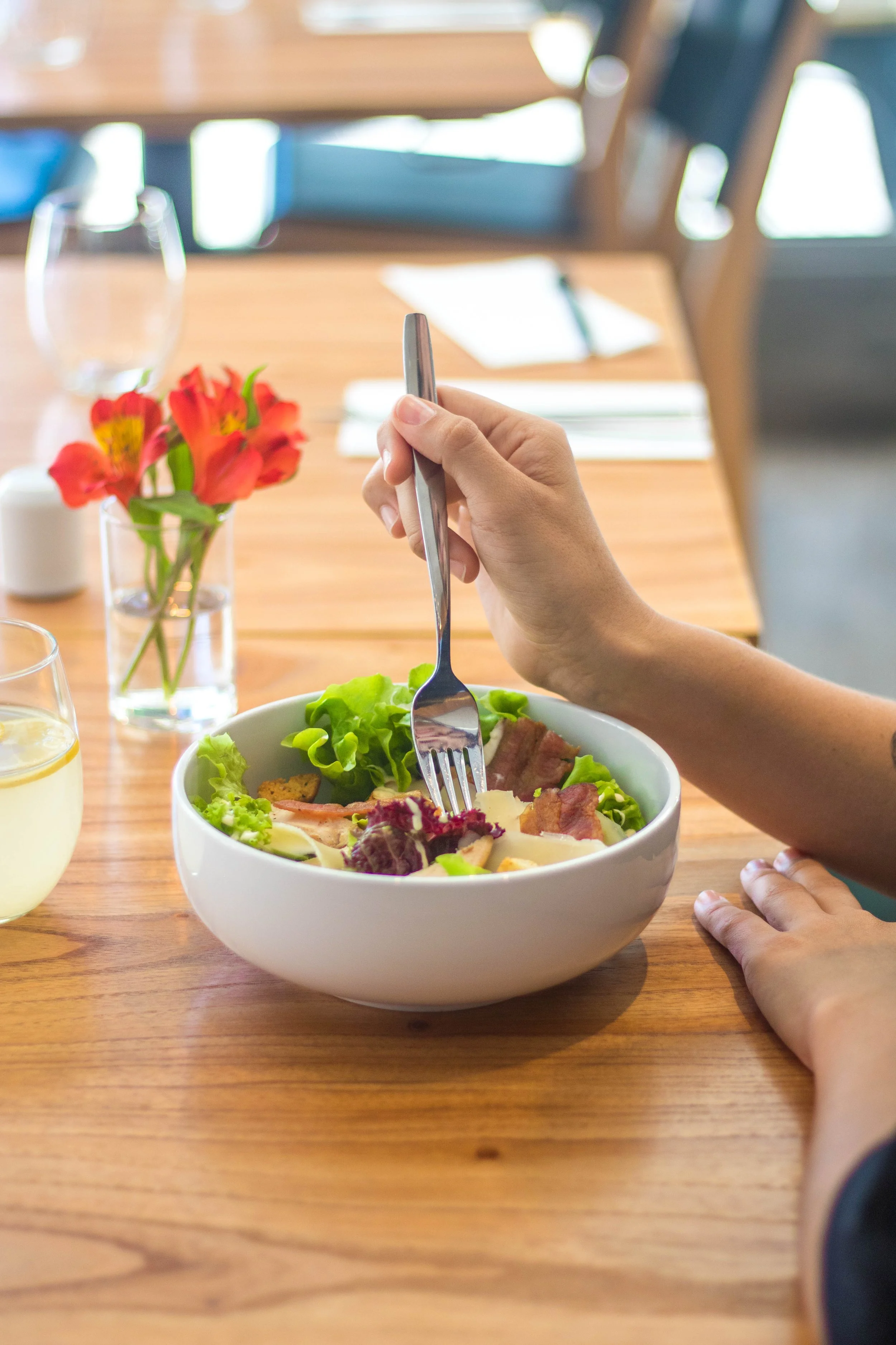 Persona poniendo comida en un tazón de ensalada en una mesa de madera, con una flor y vasos en el fondo.