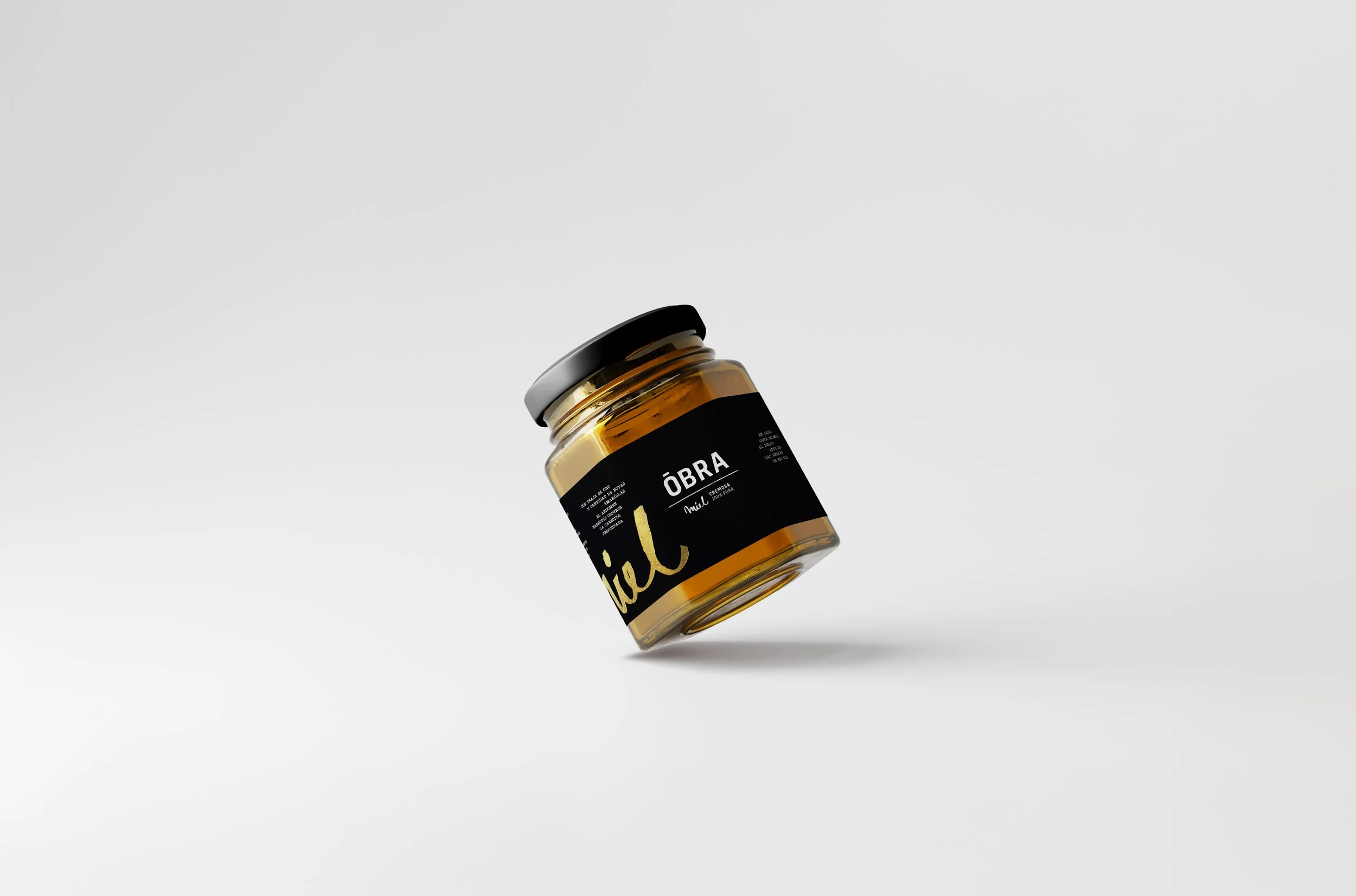 Envase de vidrio con un contenido de color dorado, etiquetado con un diseño moderno en negro y dorado que dice 'Obra' en letras grandes.