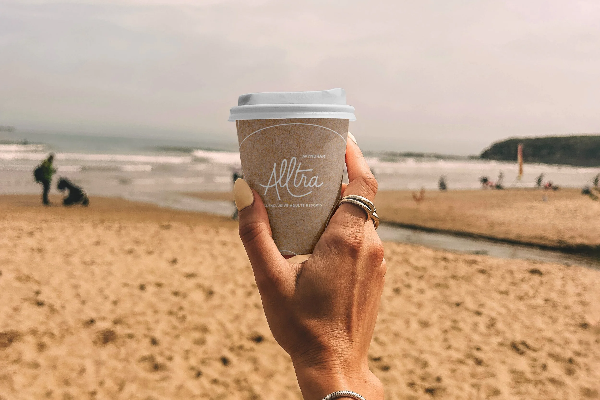 Mano sosteniendo un vaso de café con vista a la playa, con arena y olas en el fondo.
