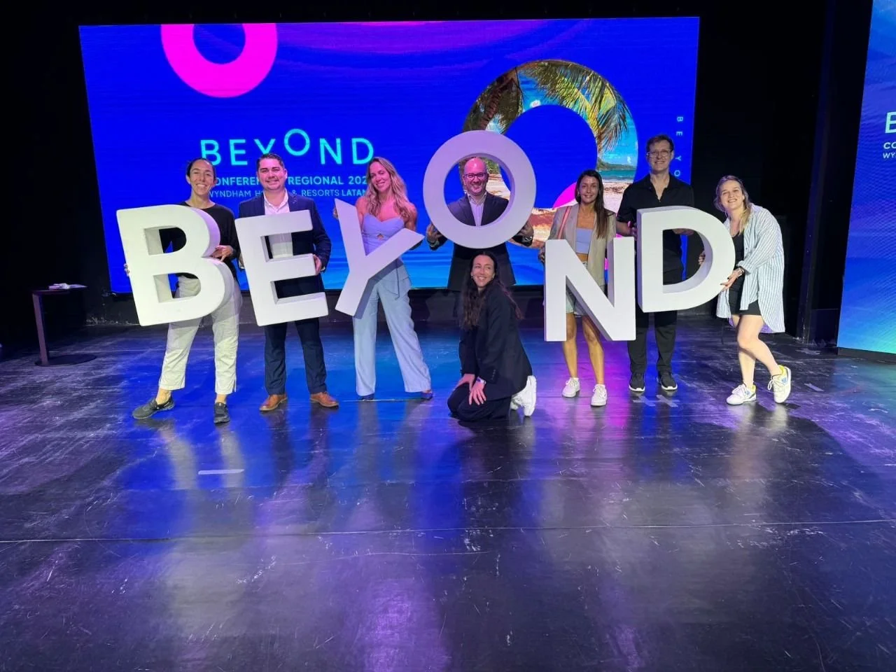 Grupo de ocho personas en escenario sosteniendo letras grandes que forman la palabra "BEYOND" en un evento con pantallas digitales de fondo que muestran un paisaje tropical y el texto "BEYOND CONFERENCIA REGIONAL 2023".