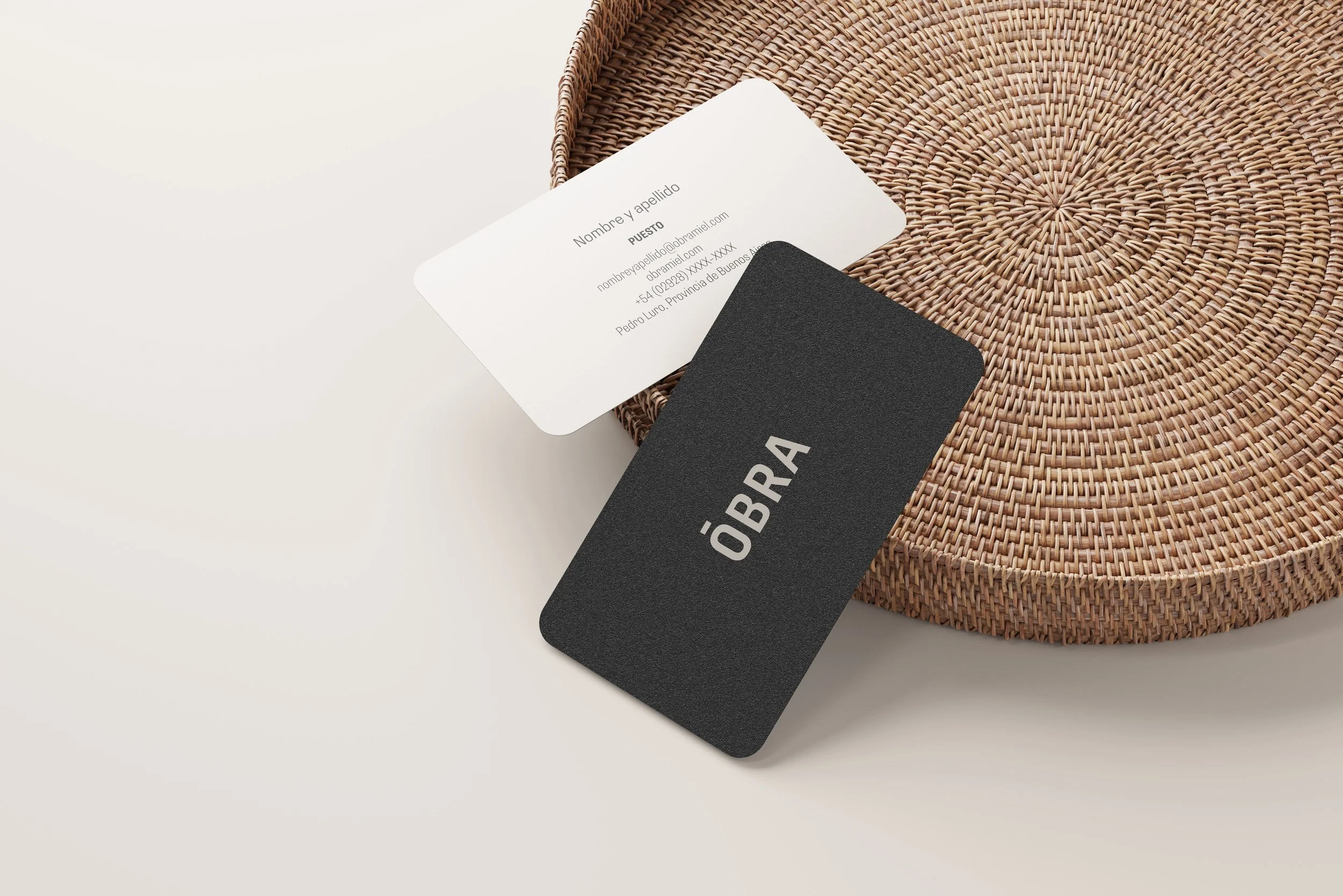 Tarjeta de presentación blanca y tarjeta negra con la palabra 'óBRA' en una superficie de mimbre redonda.