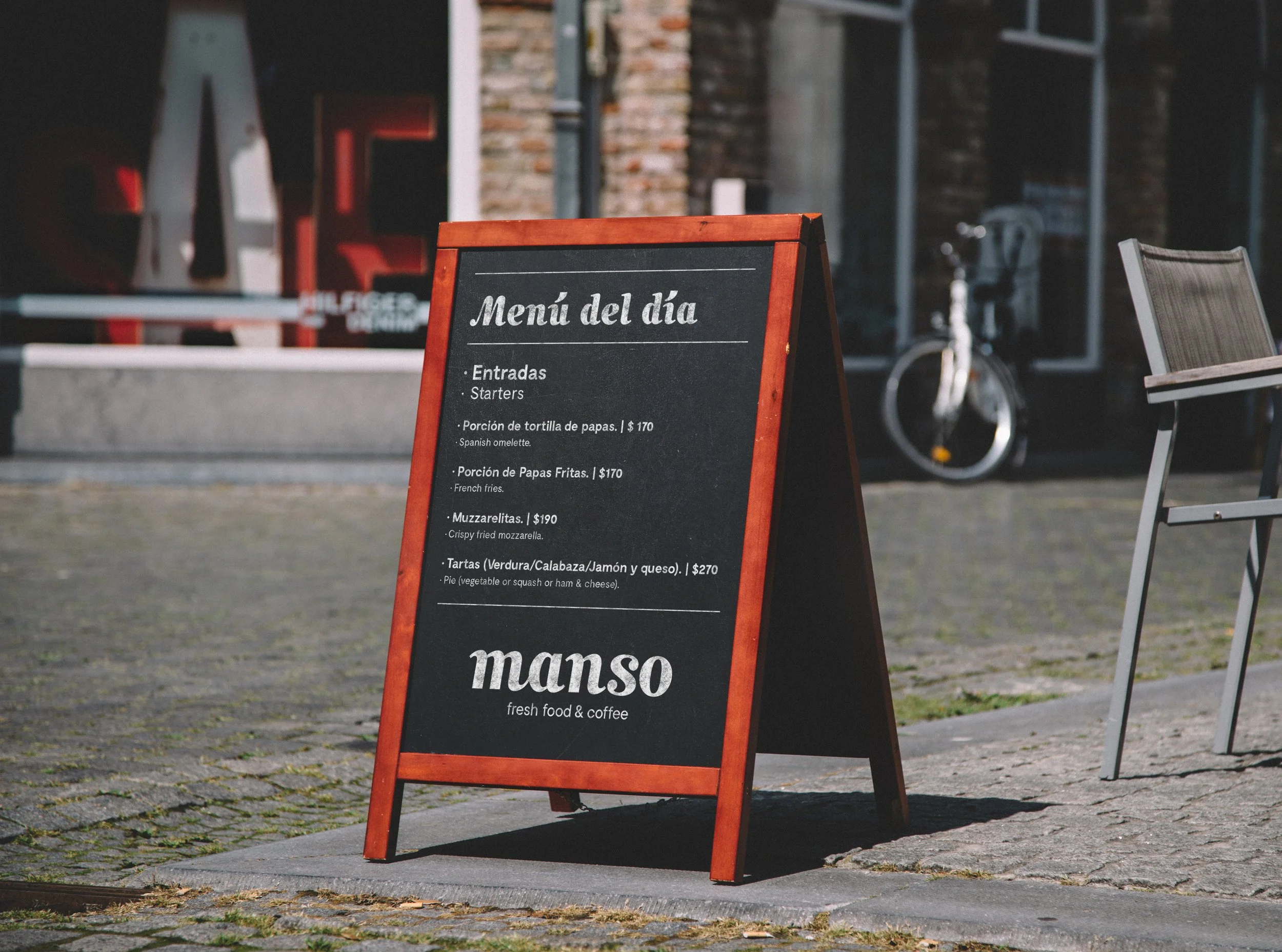 Cartel de menú del día en una cafetería con fondo de una bicicleta y un edificio, con texto en español y algunos en inglés.