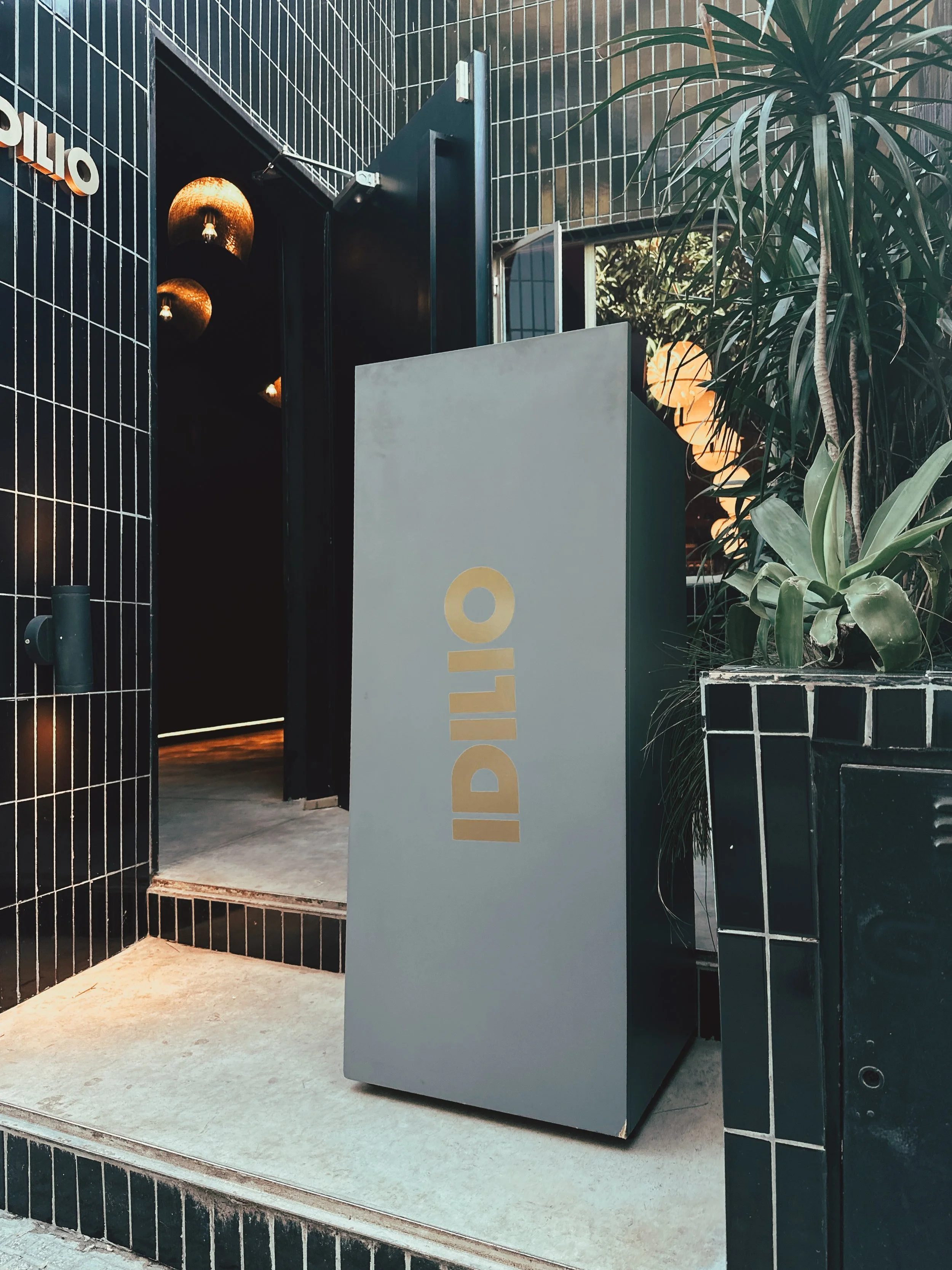 Entrada a un establecimiento llamado 'IDILIO' con un letrero gris, rodeado de paredes de azulejos negros y plantas verdes.