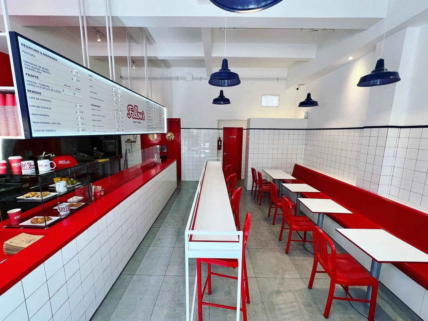 Interior de una cafetería con sillas rojas, mesas blancas, paredes blancas con azulejos, estantería con vasos y comida, cartel de menú en pantalla y luz suspendida negra.