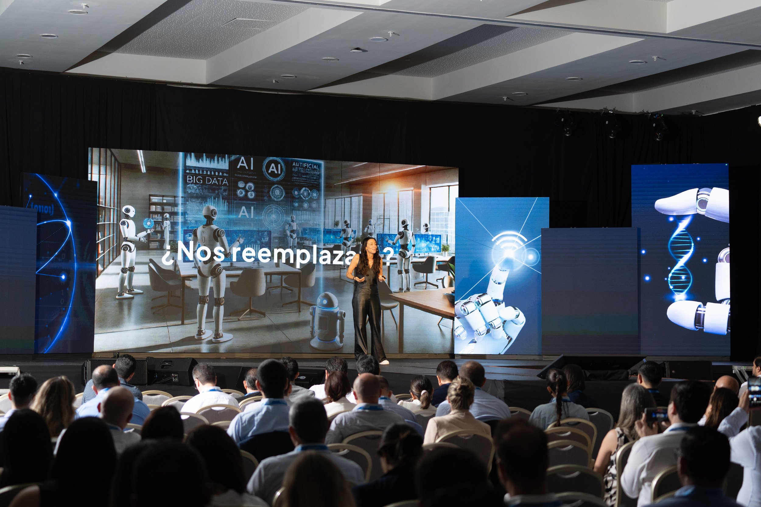 Conferencia sobre avances en inteligencia artificial en un auditorio lleno de asistentes, con una presentadora en el escenario y pantallas que muestran robots y DNA.