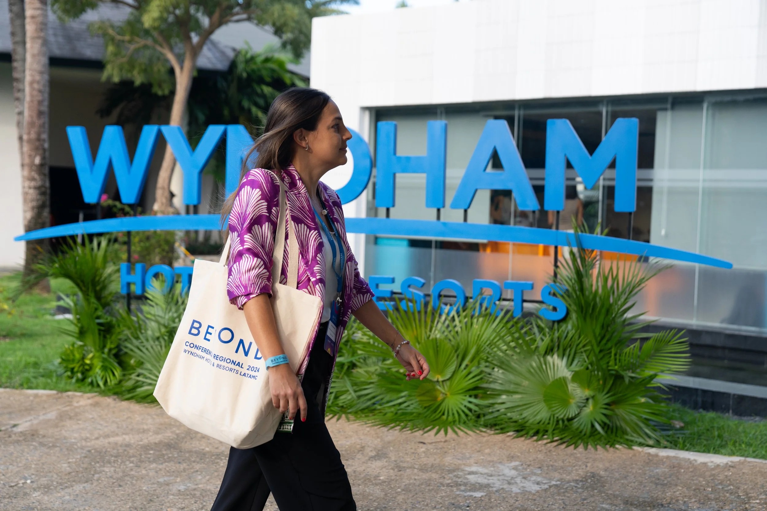 Mujer caminando frente a un letrero azul que dice "Wyndham Hotels & Resorts", llevando una bolsa de lona y una credencial en el cuello, con árboles y vegetación en el fondo.