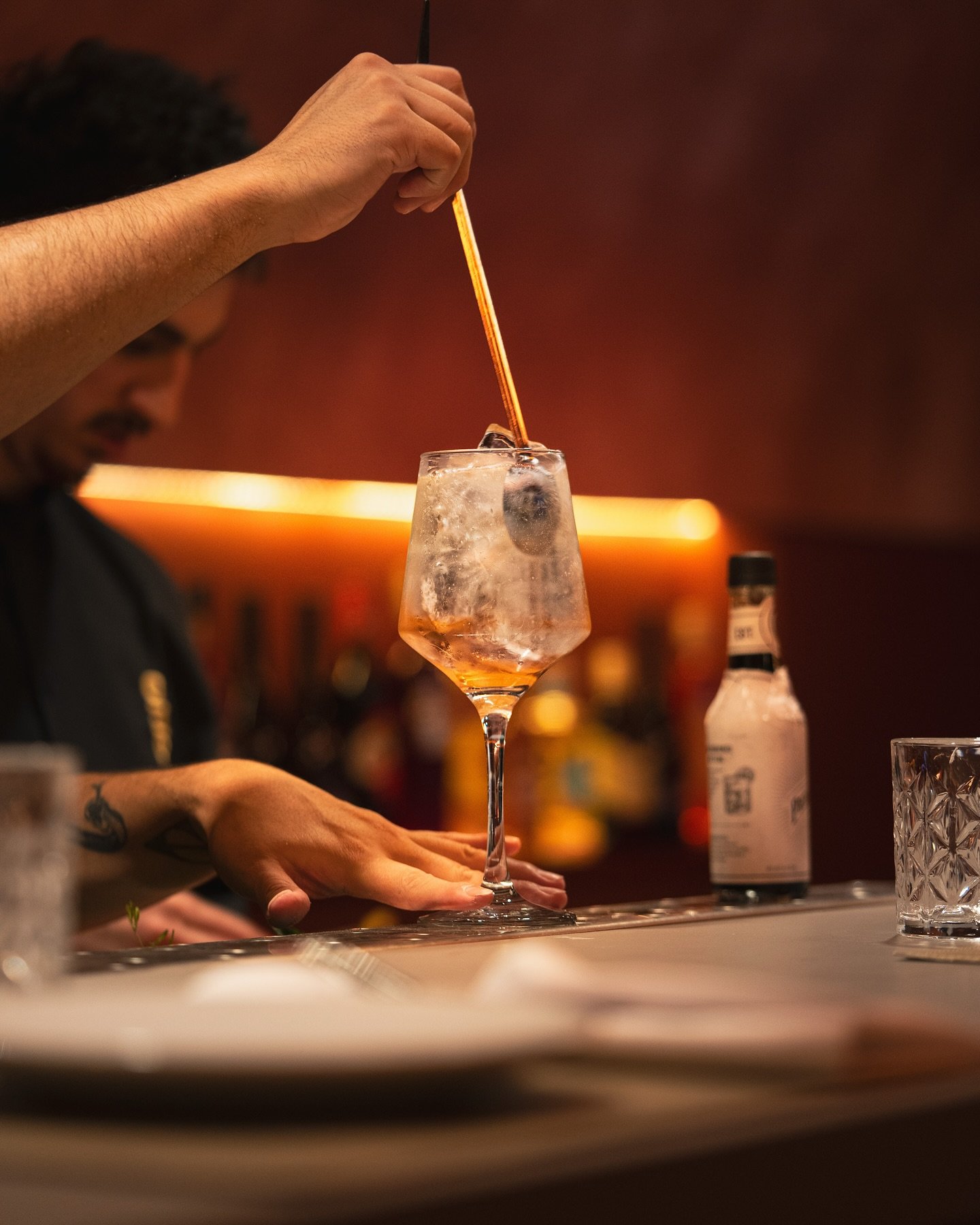 Persona mezclando una bebida en una copa en un bar con fondo oscuro y botellas de licor.