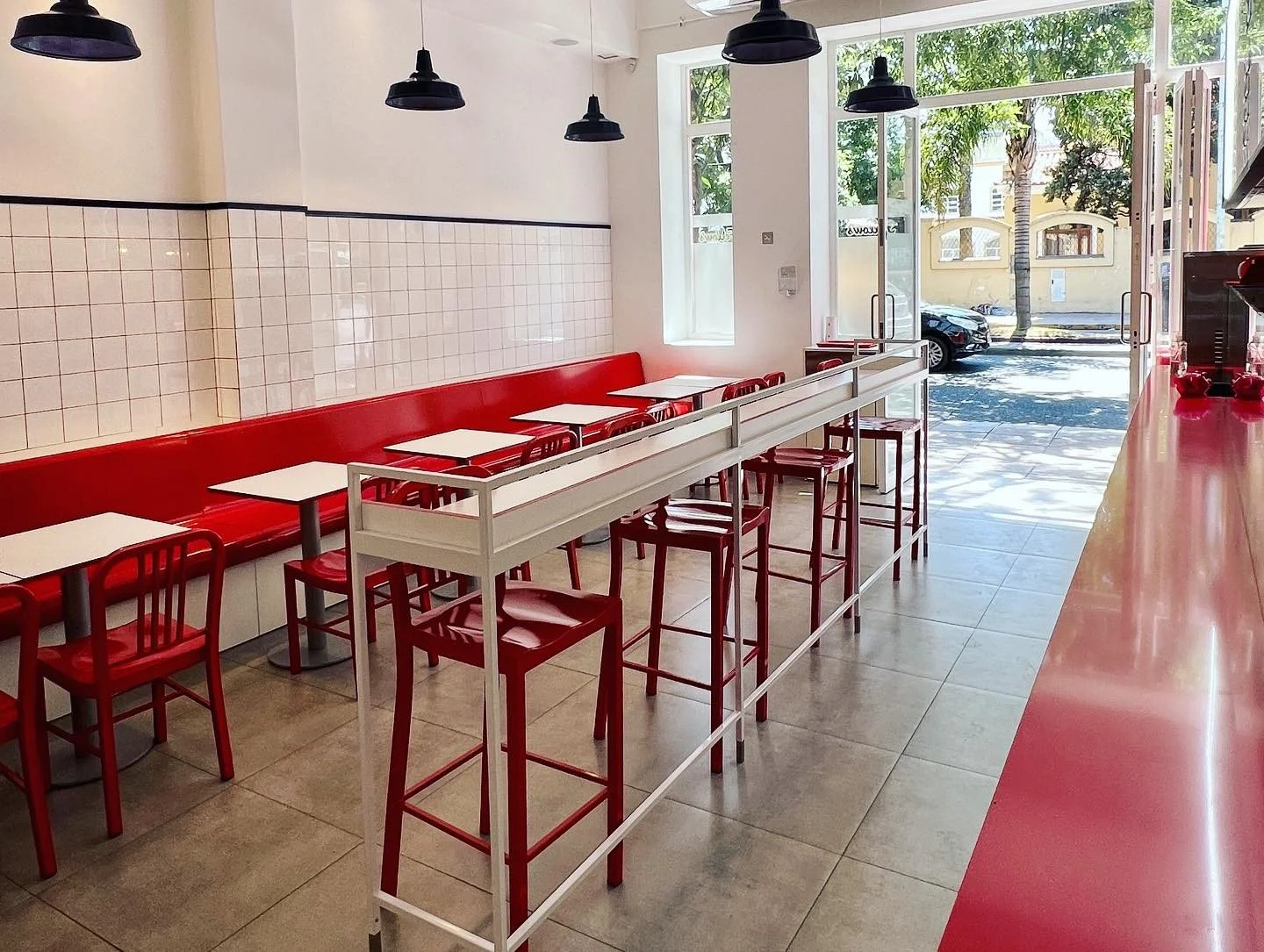 Interiores de una cafetería con sillas y mesas blancas y rojas, paredes decoradas con azulejos blancos y un gran ventanal que da hacia la calle con árboles y carros.