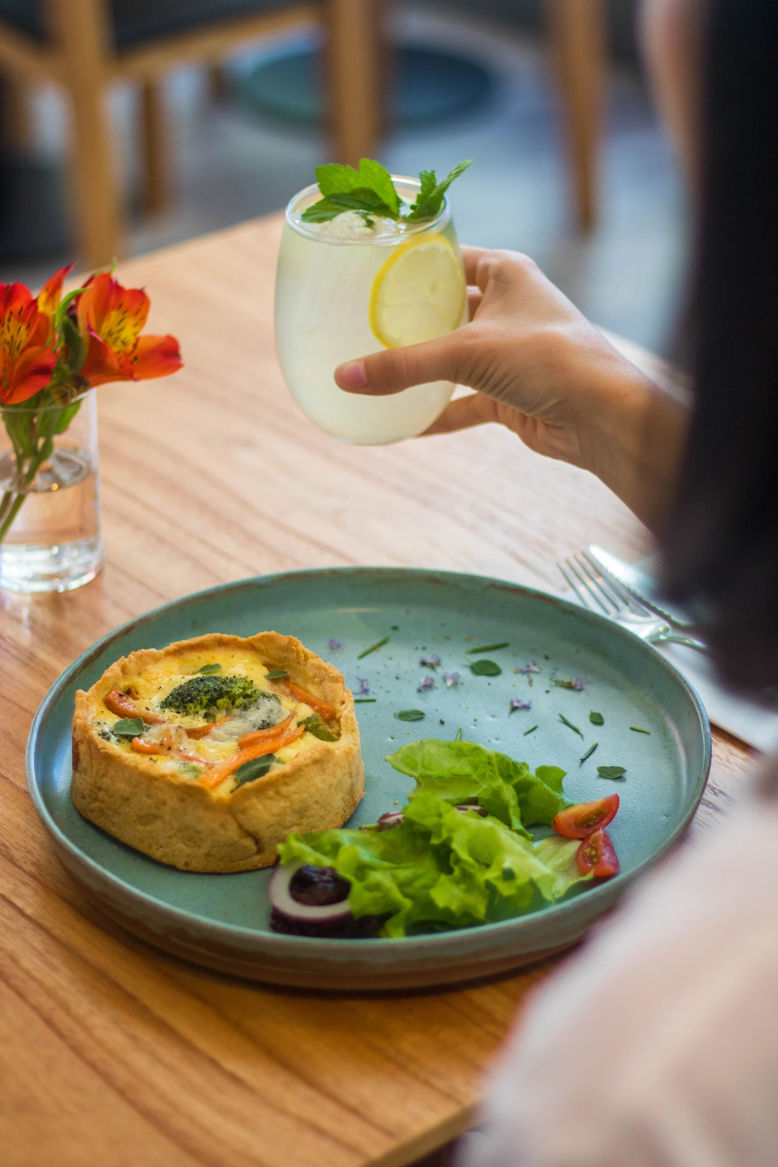 Persona sosteniendo un vaso con bebida fría decorada con rodajas de limón y hojas de menta, en un restaurante. En la mesa, plato con tarta salada, ensalada con tomates y lechuga, y jarrón con flores anaranjadas.