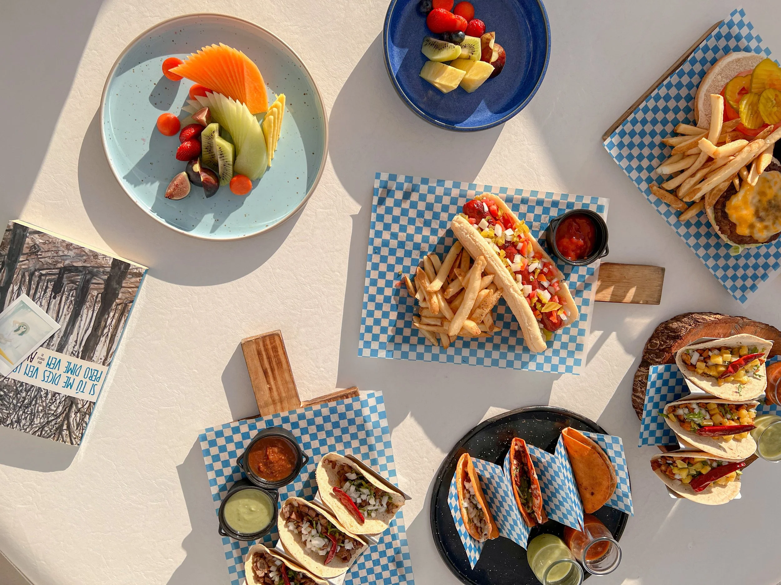 Variedad de comida y fruta en platos y bandejas en una mesa, incluyendo tacos, hot dogs y ensalada de frutas.