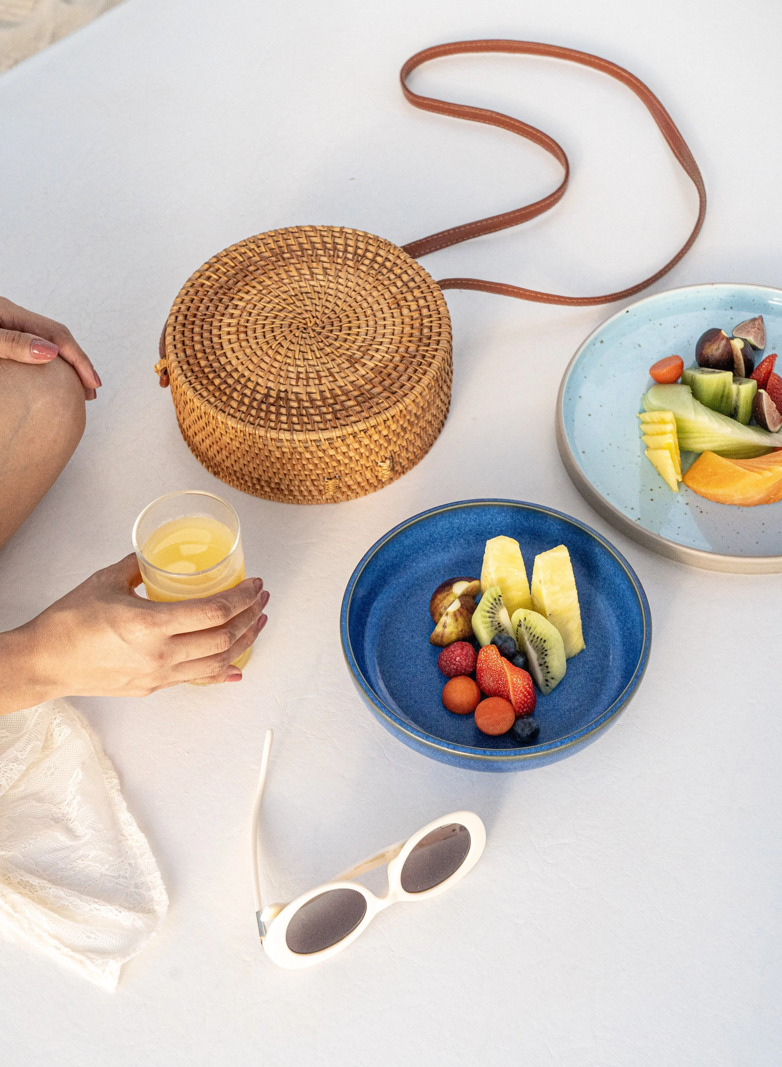 Vasos con jugo y fruta, varios platos con frutas, gafas de sol blancas, bolso de mimbre, mano con vaso con jugo, paño de tela