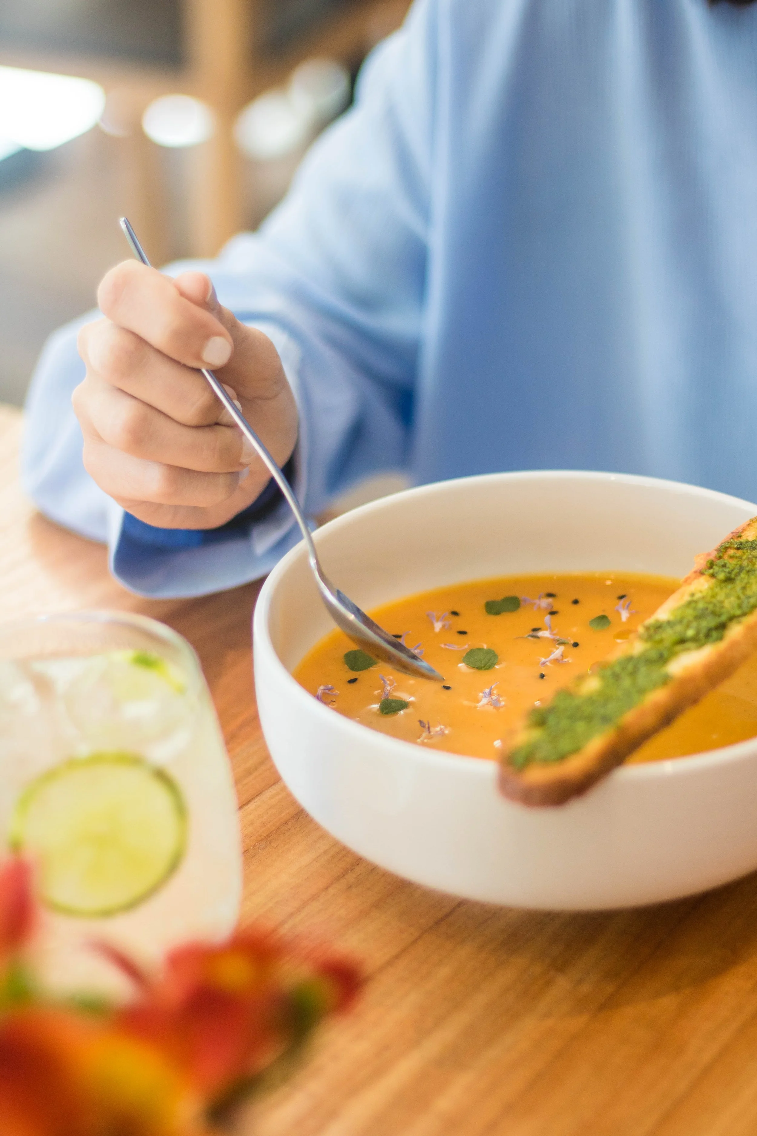 Persona sirviendo una sopa de color naranja en un bol blanco, decorada con flores y hierbas, con una tostada con guacamole o aguacate al lado, en una mesa de madera.