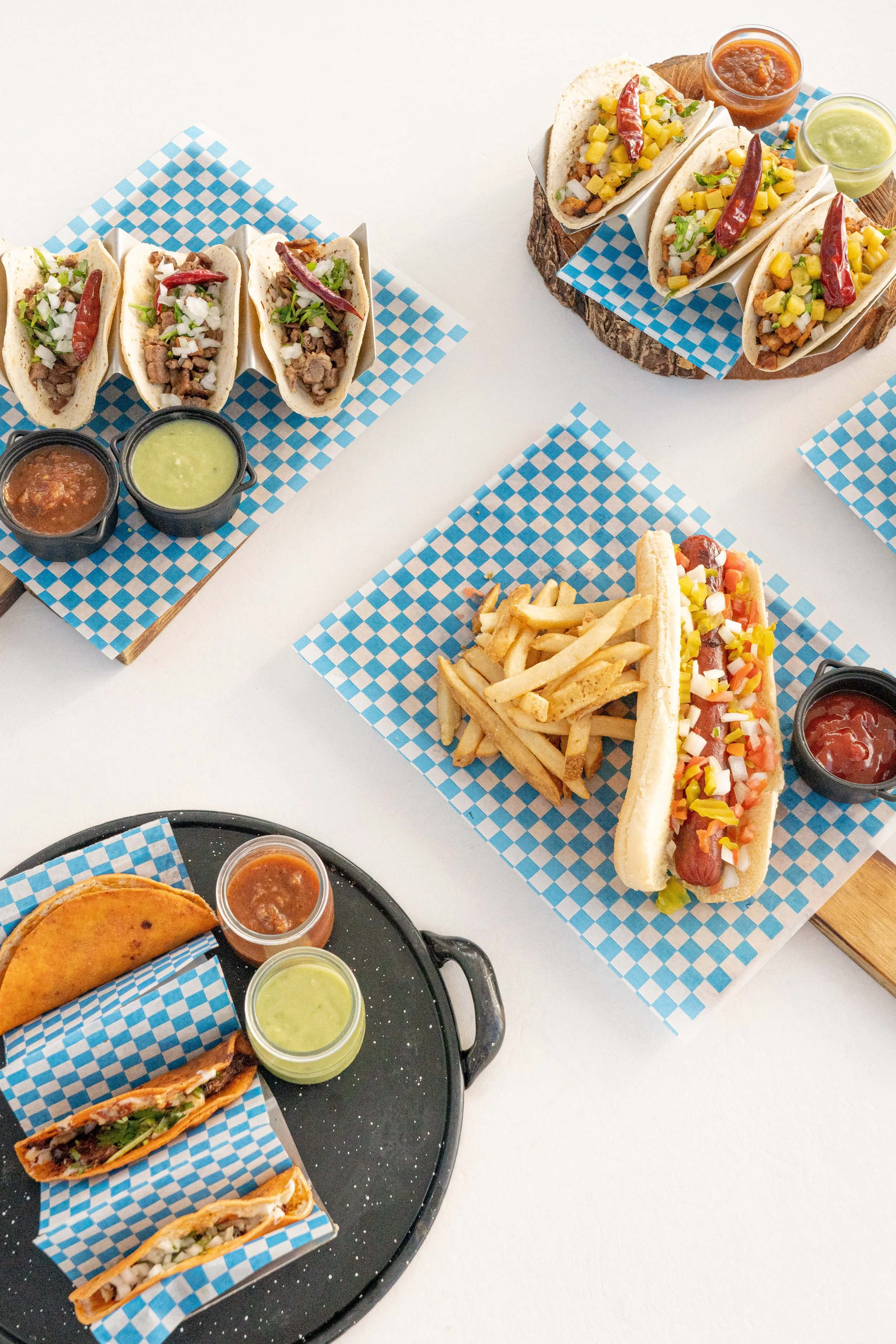 Mesa con tacos, papas fritas y salsas variadas en diferentes platos y fondos blancos.