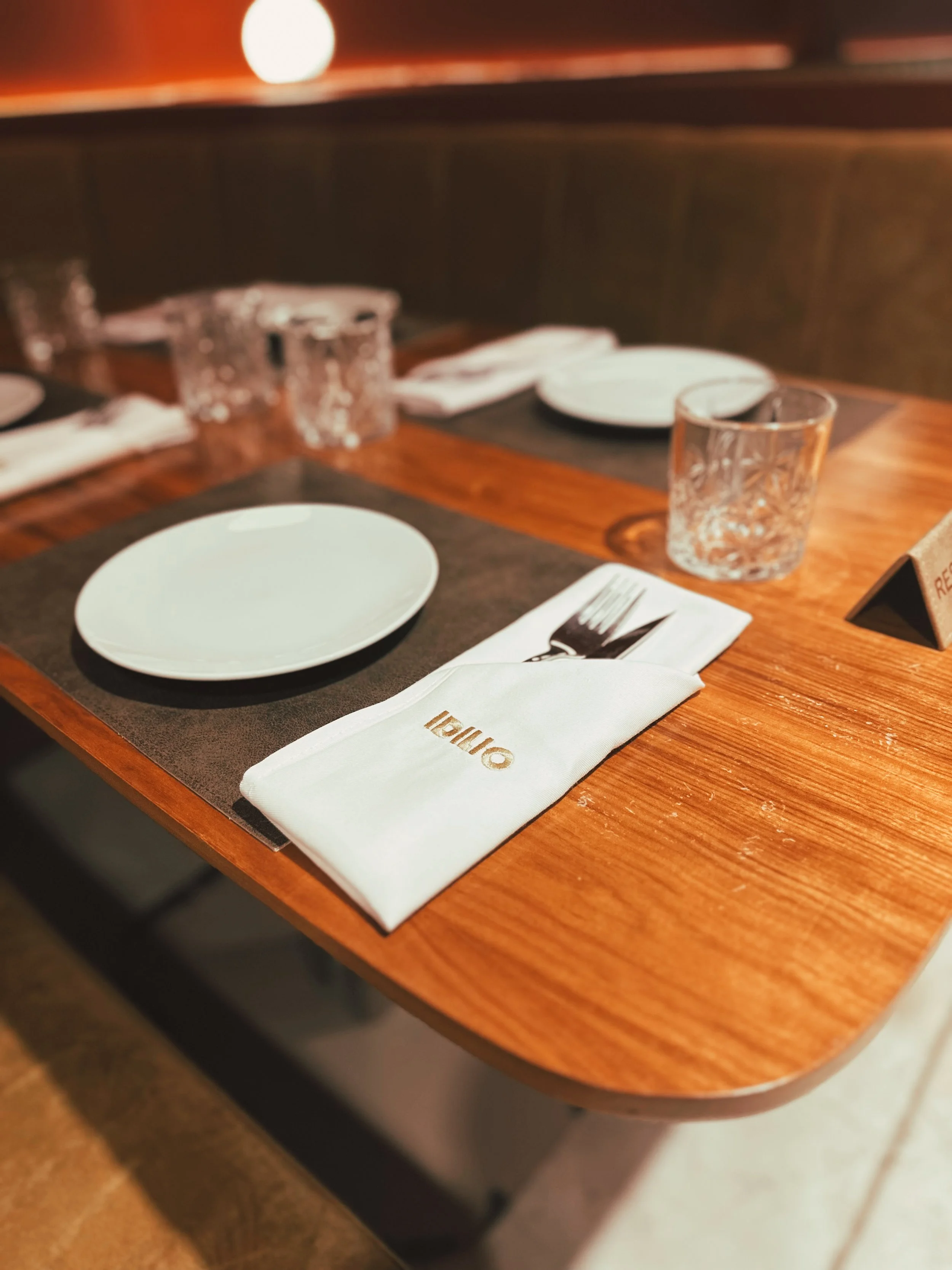 Mesa de restaurante con platos blancos, vasos de agua cortos, cubiertos envueltos en servilletas blancas con la inscripción 'Oino', y un cartel de reservación.