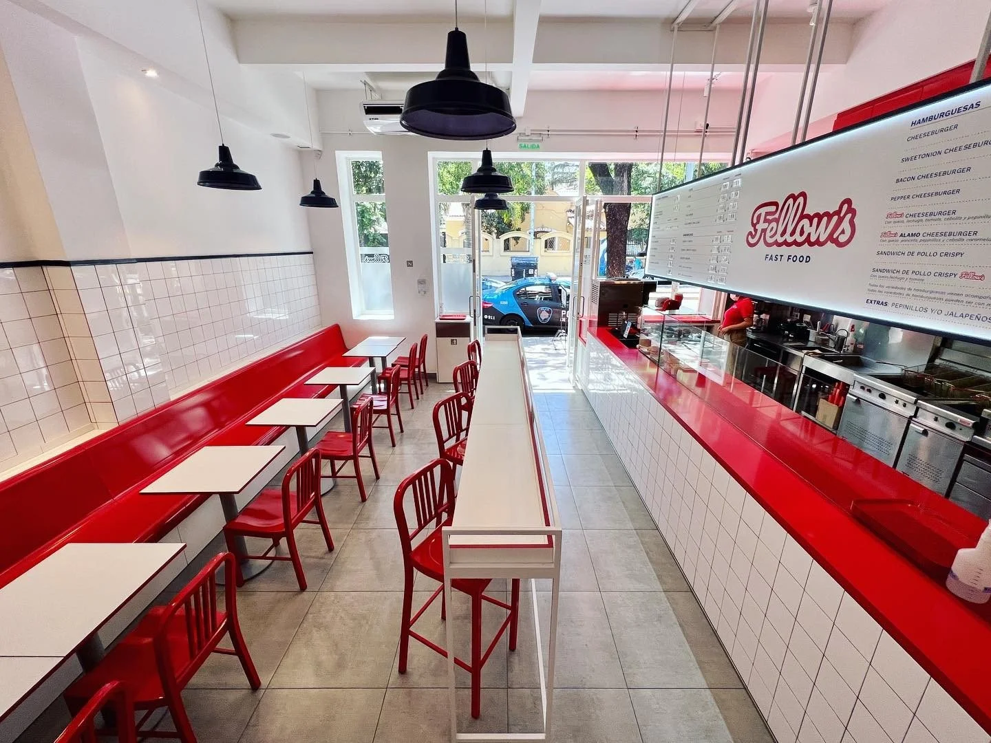 Interior de una pizzería con mesas blancas y sillas rojas, paredes blancas con azulejos en la parte inferior, y una barra de servicio también en rojo. Frente a la barra, un menú digital de color blanco con letras rojas, y una ventana que deja ver un 