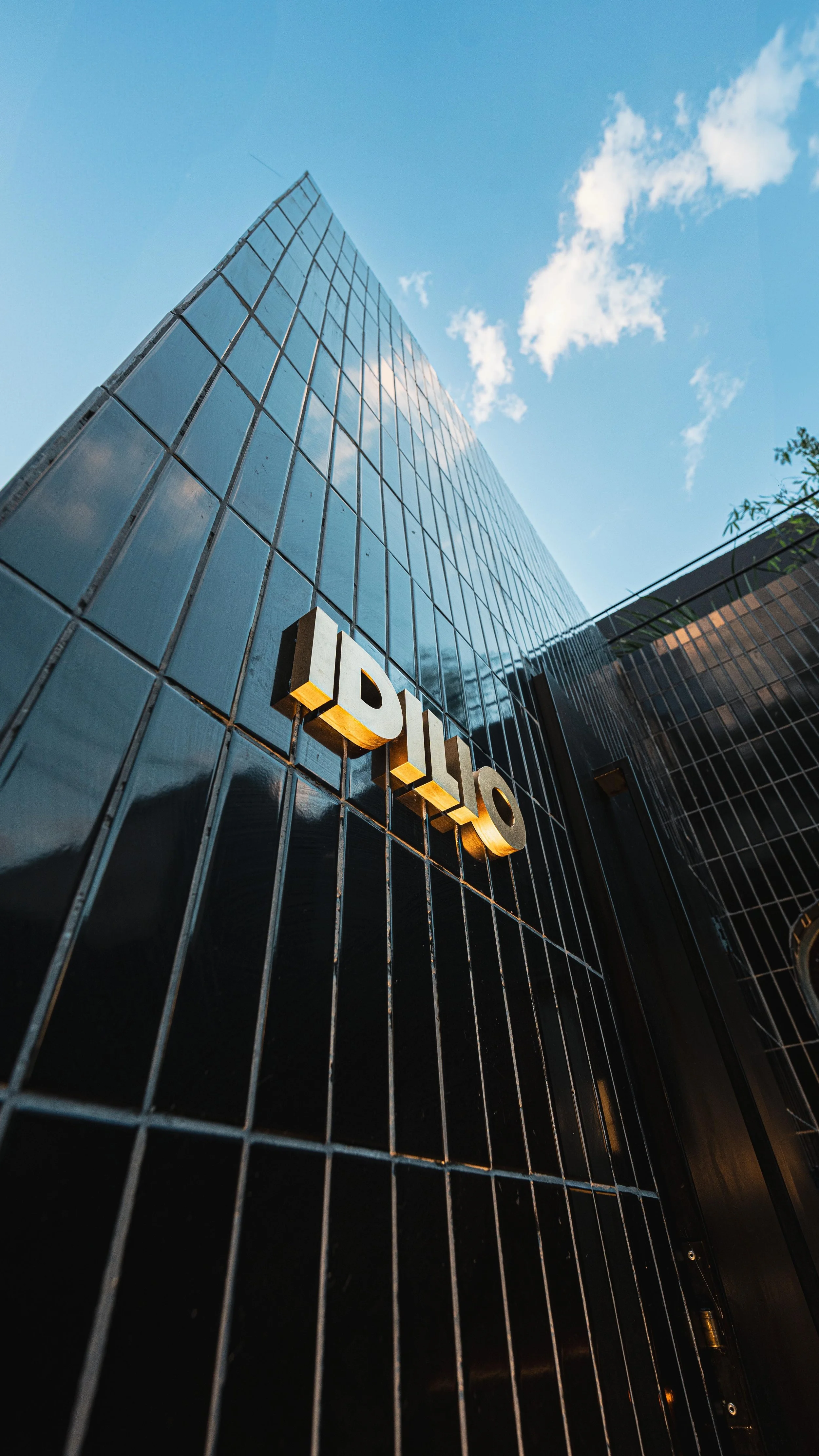 Edificio moderno de cristal con cartel de IDIILO en la fachada, vista desde abajo hacia el cielo azul con nubes.