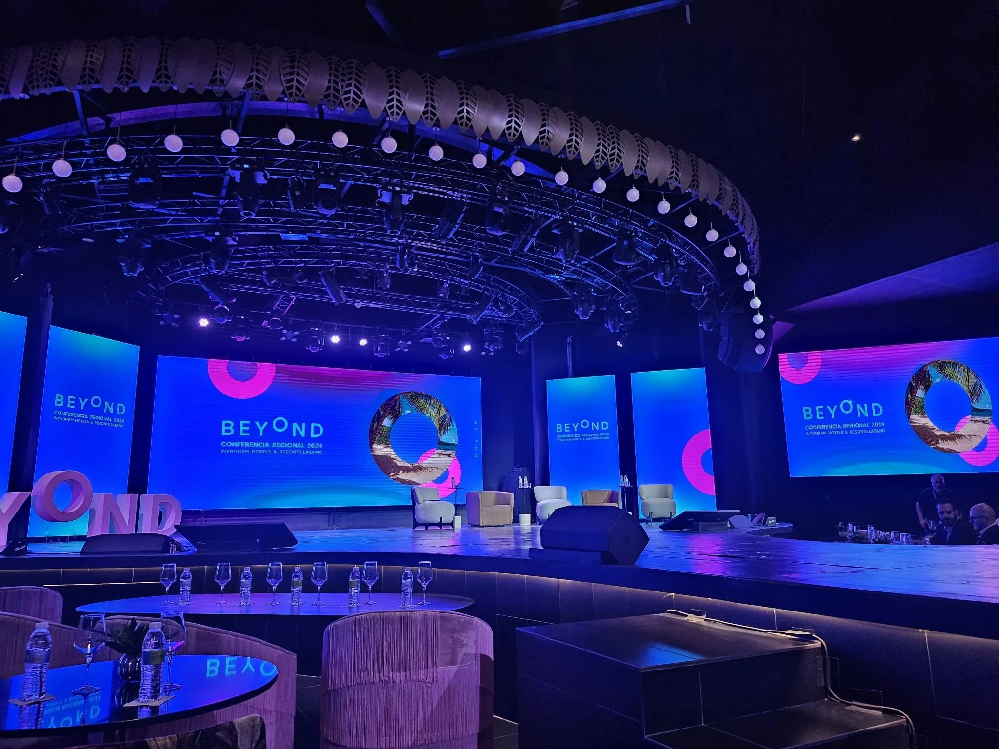 Escenario de una conferencia con pantallas digitales azules y rosa que muestran el texto 'BEYOND' y un logo circular, con sillas y mesas preparadas para la audiencia