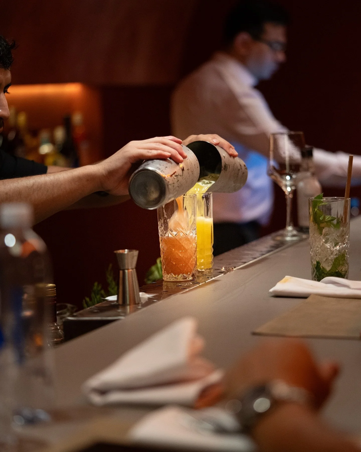 Barista sirviendo cócteles en un bar elegantes, con varias bebidas en copas decoradas, mientras otro bartender trabaja en el fondo.