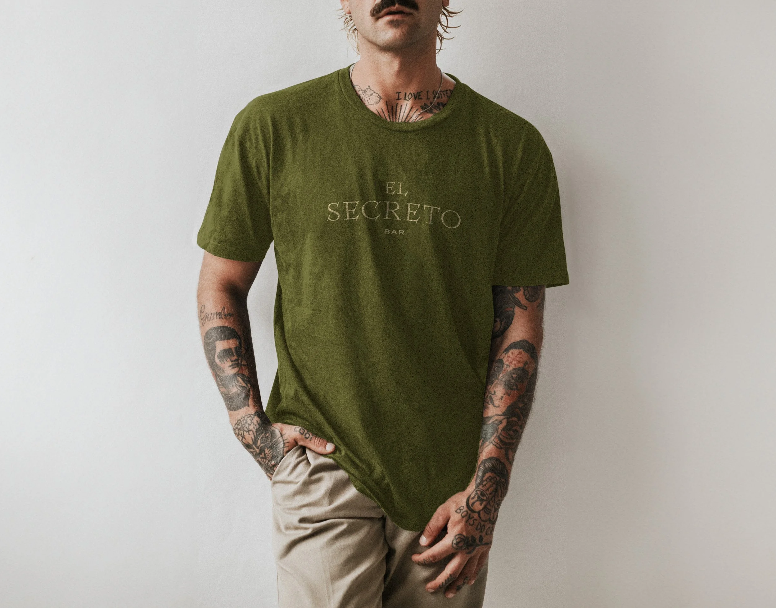 Hombre con tatuajes en brazos y cuello, lleva una camiseta verde con la inscripción 'El Secreto Bar', y pantalones beige, de pie contra una pared blanca.