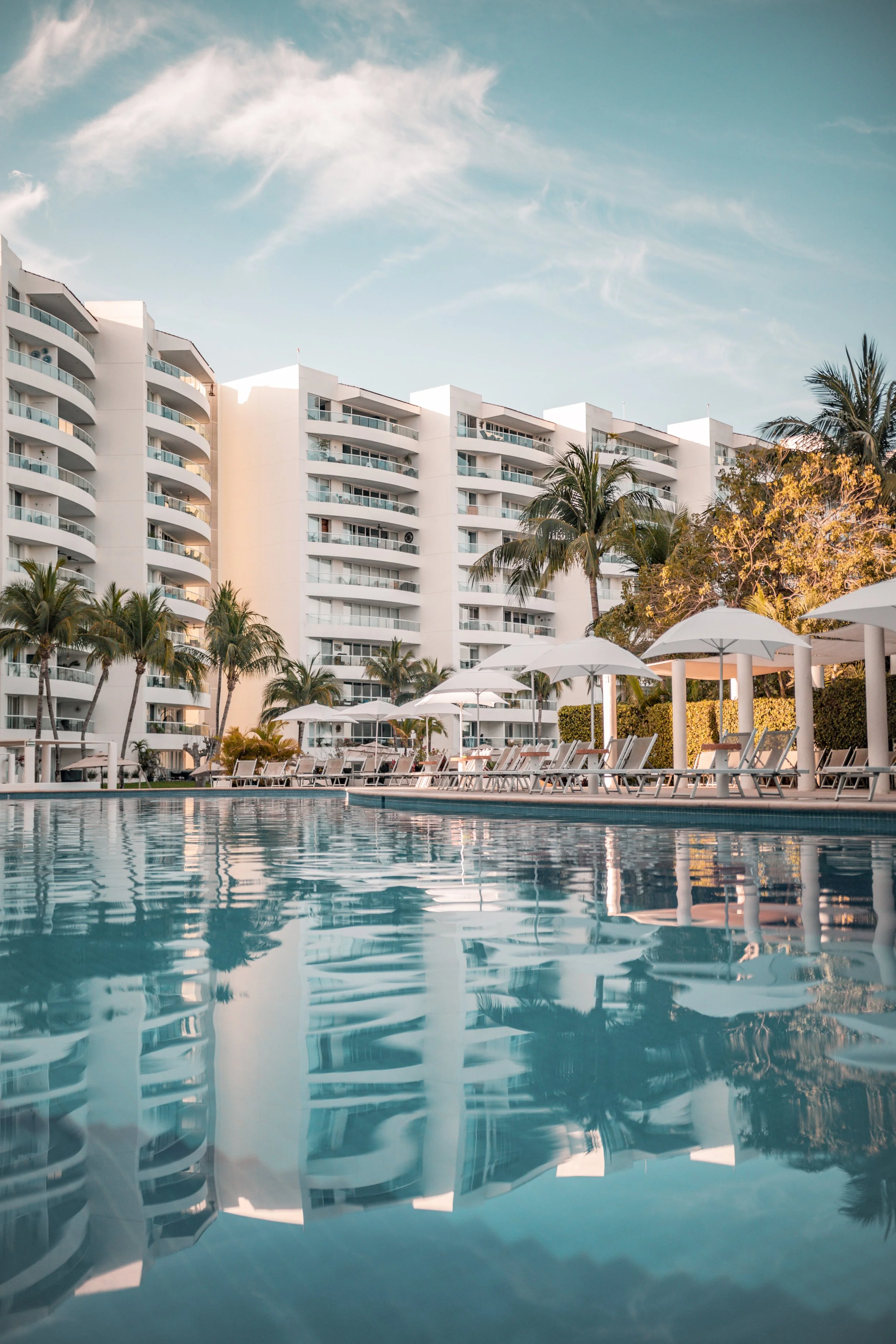 Piscina en un hotel con sillas y sombrillas blancas, edificios altos de apartamentos blancos, palmeras y un cielo con nubes finas.