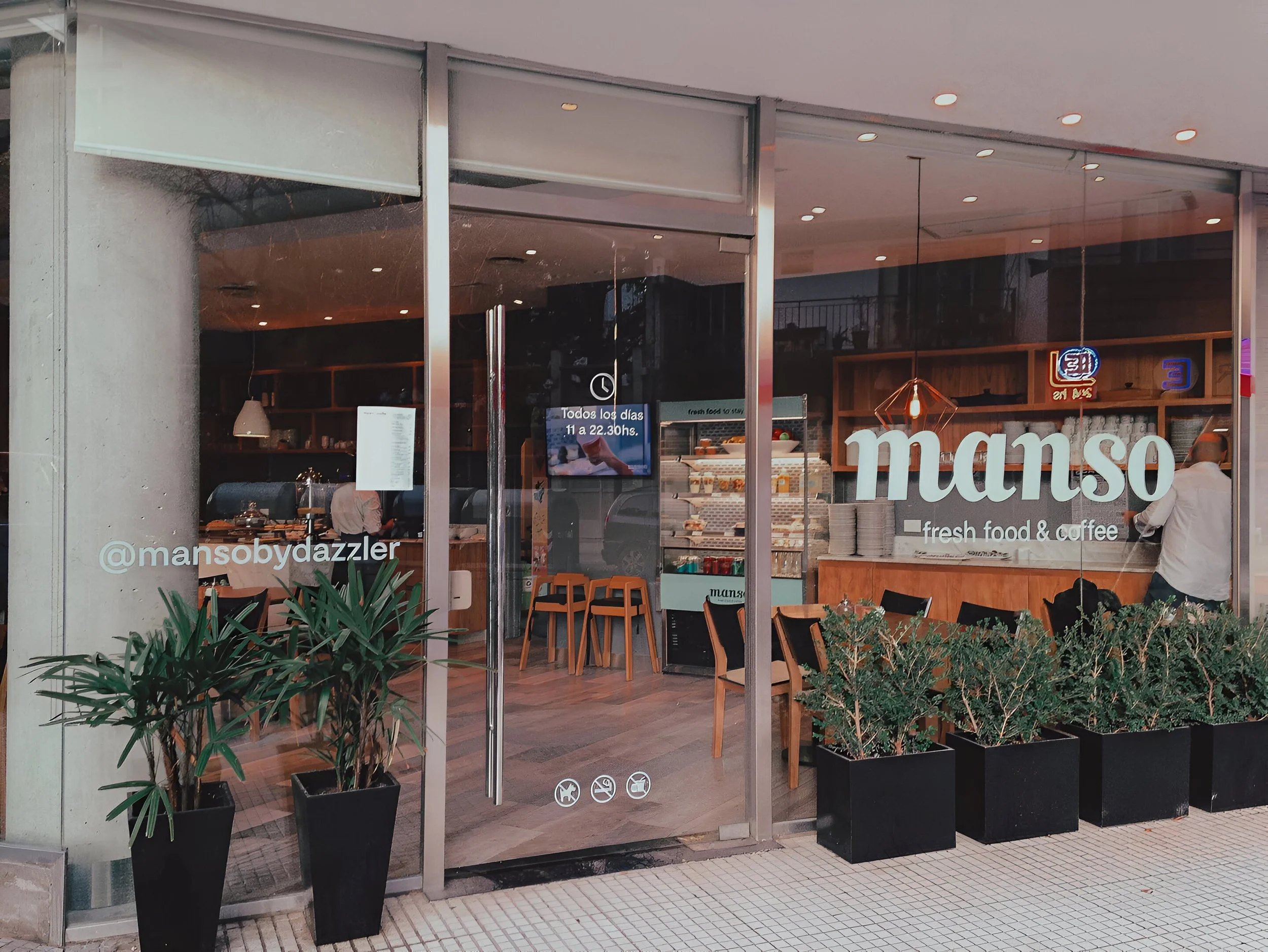 Fachada de un café llamado 'Manso' que ofrece comida fresca y café, con plantas en macetas en la entrada, ventanas de vidrio y decoraciones modernas en el interior.