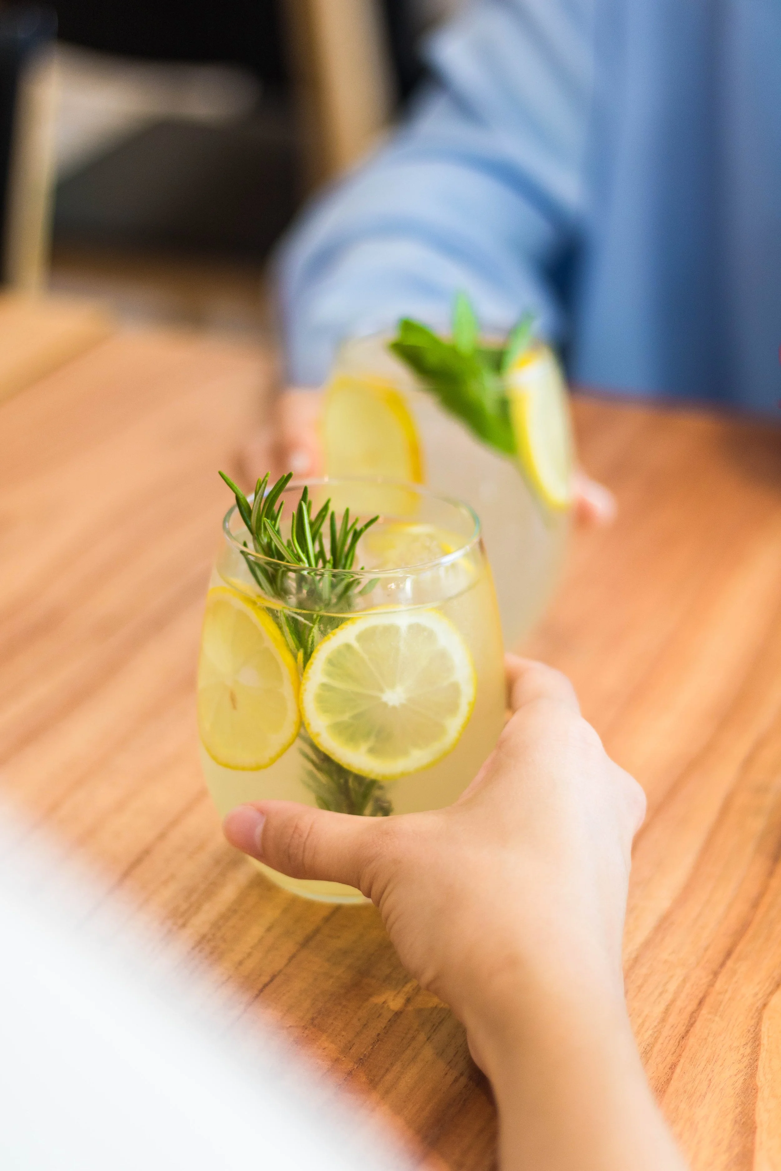 Dos vasos con bebida de limón y hierbas, uno en primer plano y otro en el fondo, en una mesa de madera.