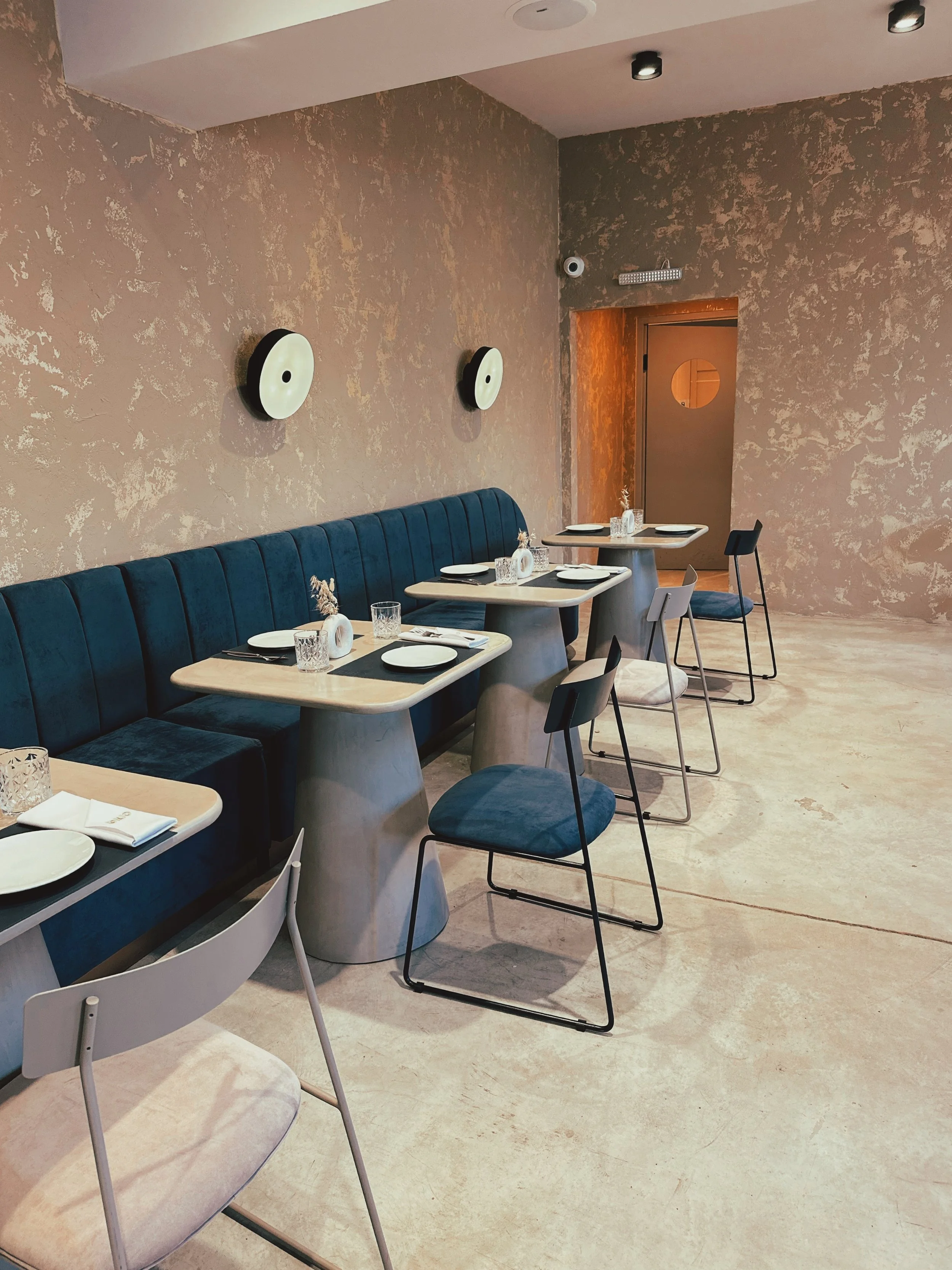 Interior de un restaurante moderno con paredes de textura beige, mesas con mantel negro y platos blancos, sillas modernas y decoración minimalista.