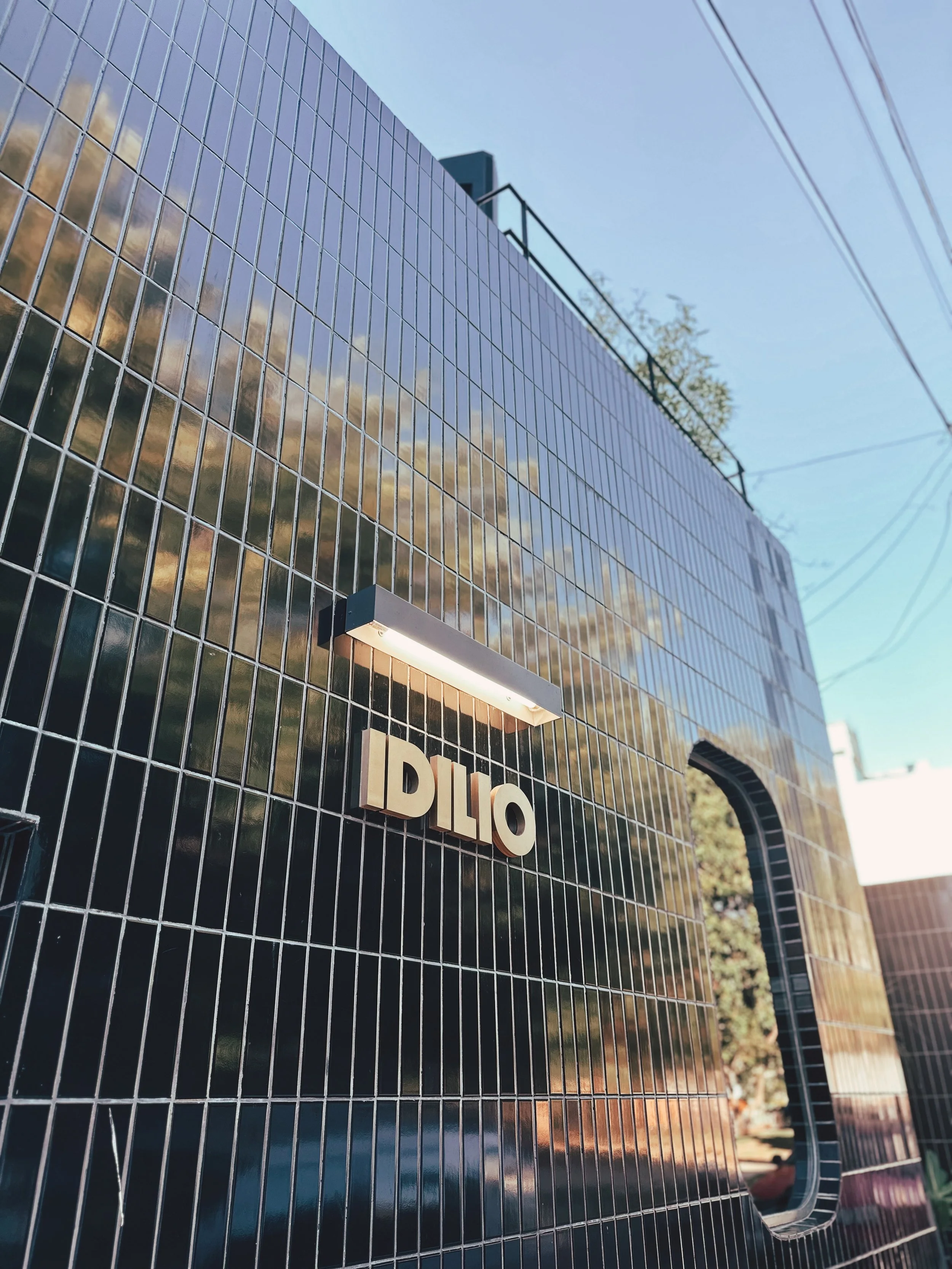 Fachada de una estructura moderna con azulejos negros y un letrero que dice 'IDILIO' con luz encima, y un arco en la esquina derecha.