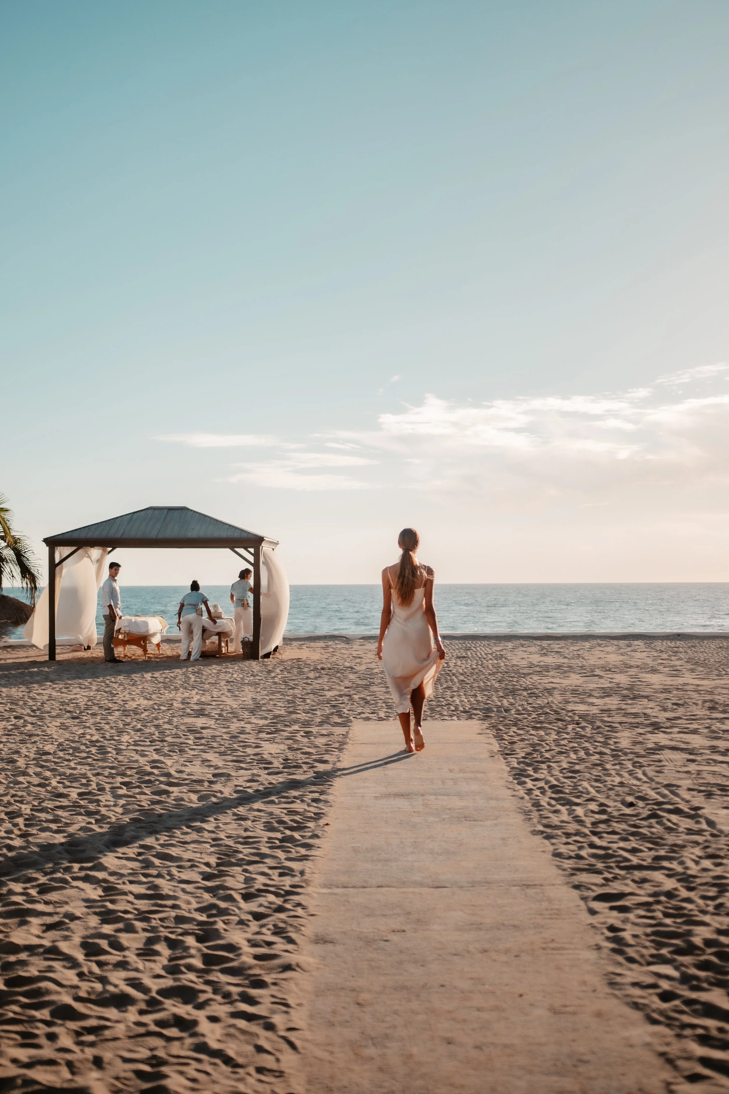 Una mujer caminando hacia una ceremonia en la playa, con el mar y el cielo al fondo, y un altar en la arena a la izquierda