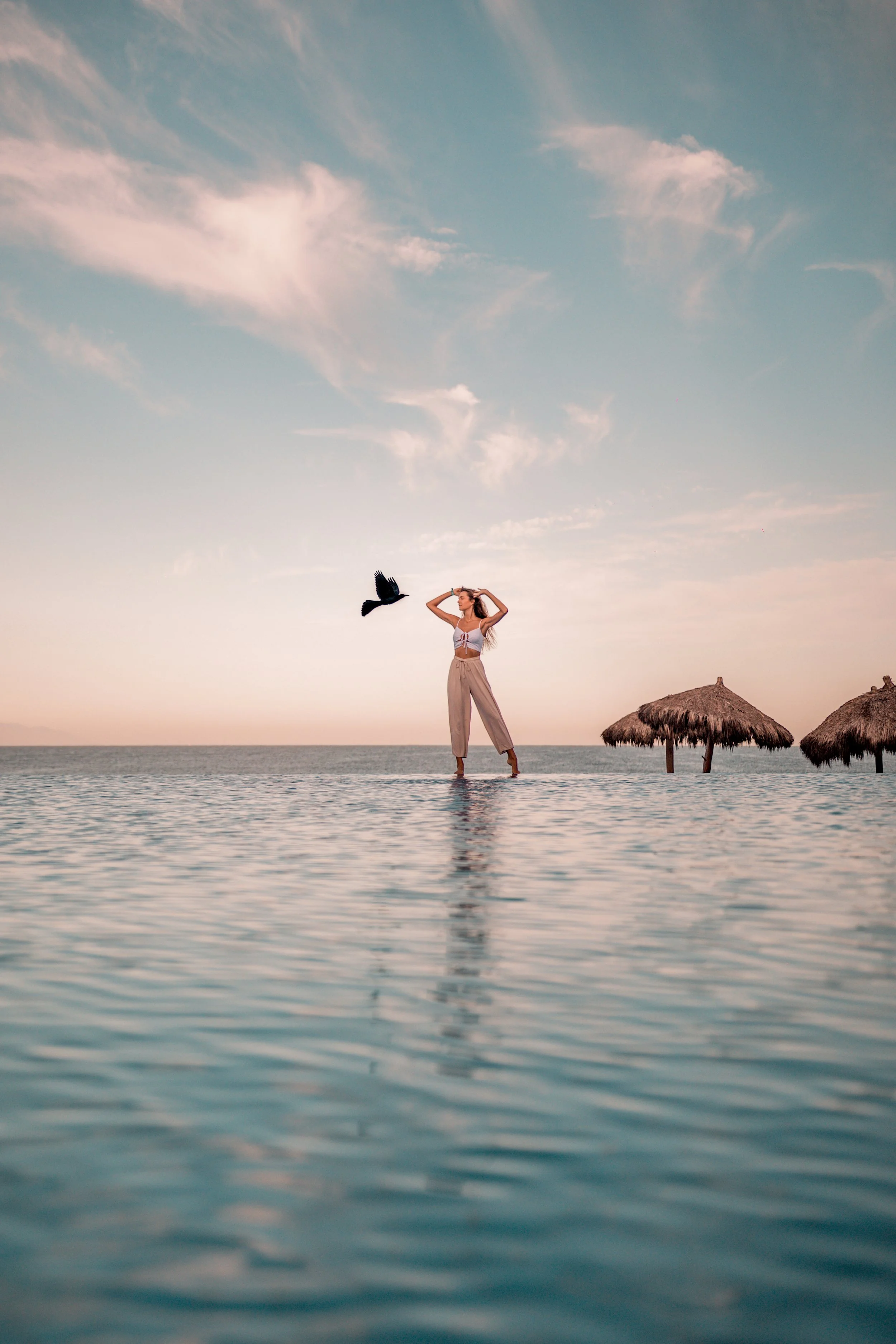 Mujer de pie en piscina en la playa con sombrillas de paja y cielo despejado.
