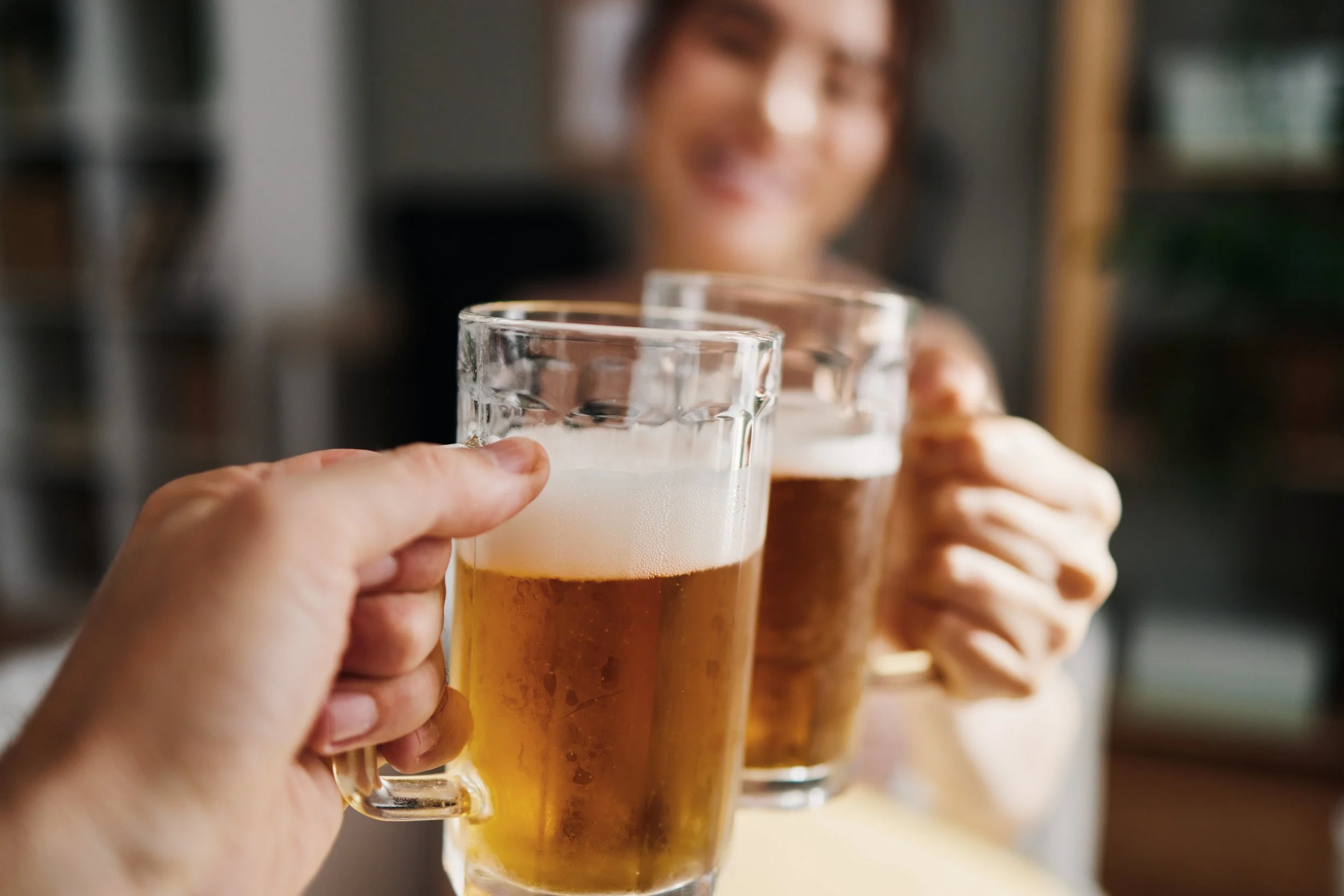 Dos personas brindando con vasos de cerveza en un ambiente acogedor, una de ellas sonriendo al fondo.