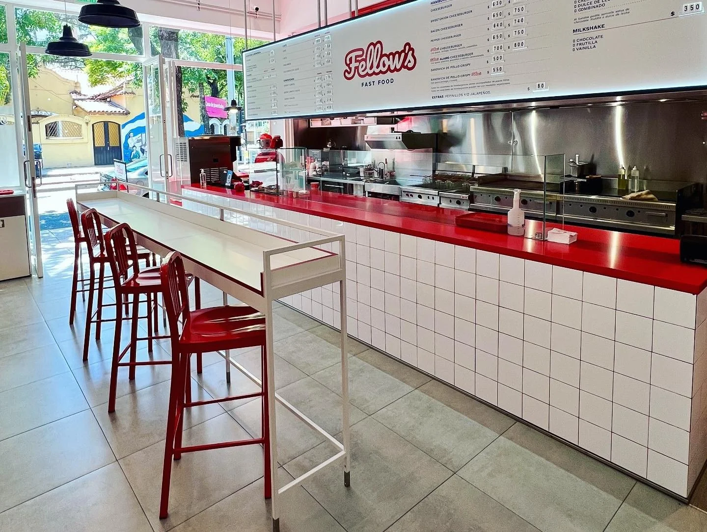 Interiores de un restaurante de comida rápida con barra de cocina, menú en la pared y sillas rojas junto a una mesa larga blanca. Entrada de vidrio con vista a la calle con árboles y casas.