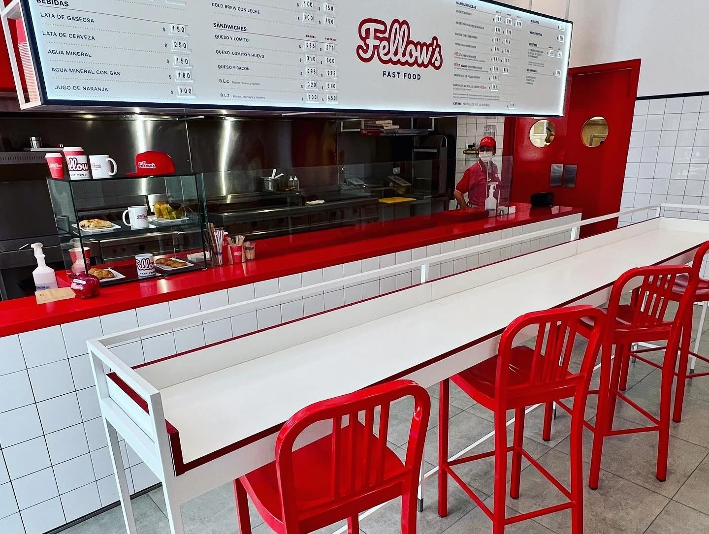 Interiores de un restaurante de comida rápida con mesas blancas y sillas rojas, mostrador con alimentos y bebidas, y un empleado con mascarilla en el fondo. Incluye cartel con menú en la parte superior.