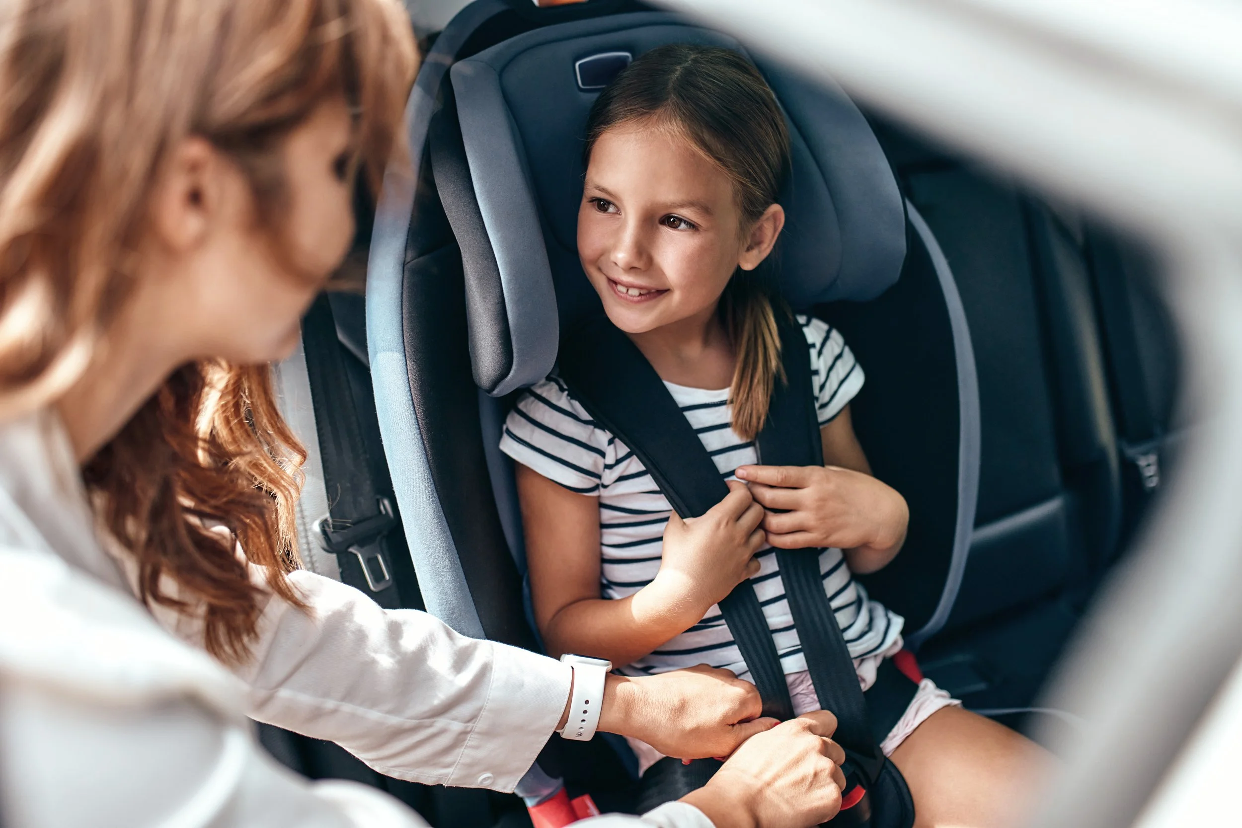 Una niña en una silla de auto ajustándose el cinturón de seguridad, mientras una mujer le ayuda, en el interior de un vehículo.
