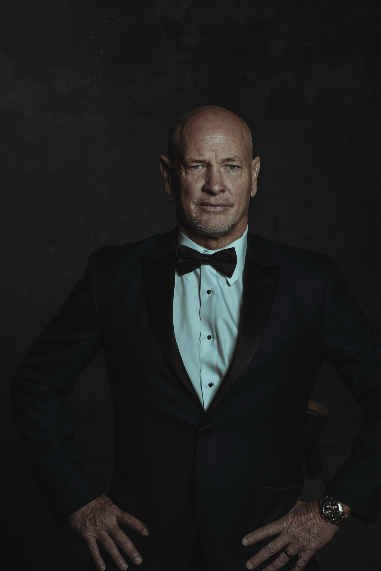 An older man with a bald head and gray goatee wearing a black tuxedo with a white shirt and black bow tie stands against a dark background. He has a confident expression, with hands resting on his hips, one of which shows a gold watch on his wrist.