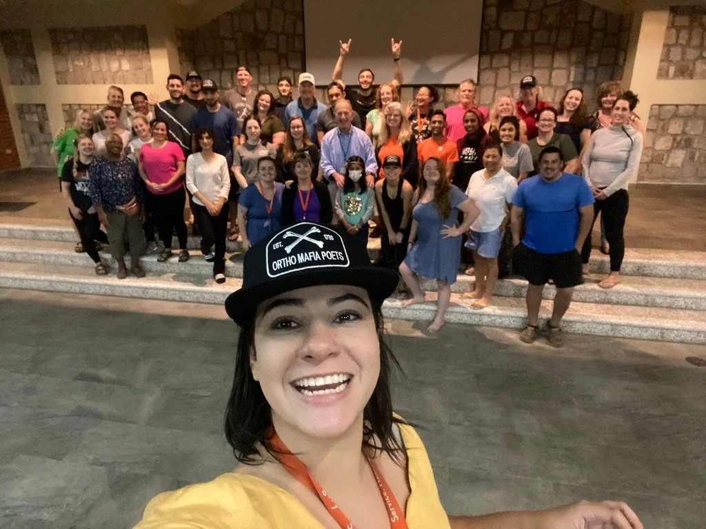 A cheerful woman wearing a black cap with 'ORTHO MAFIA POETS' written on it, taking a selfie in front of a large group of diverse people posing on steps in a spacious room with stone walls.