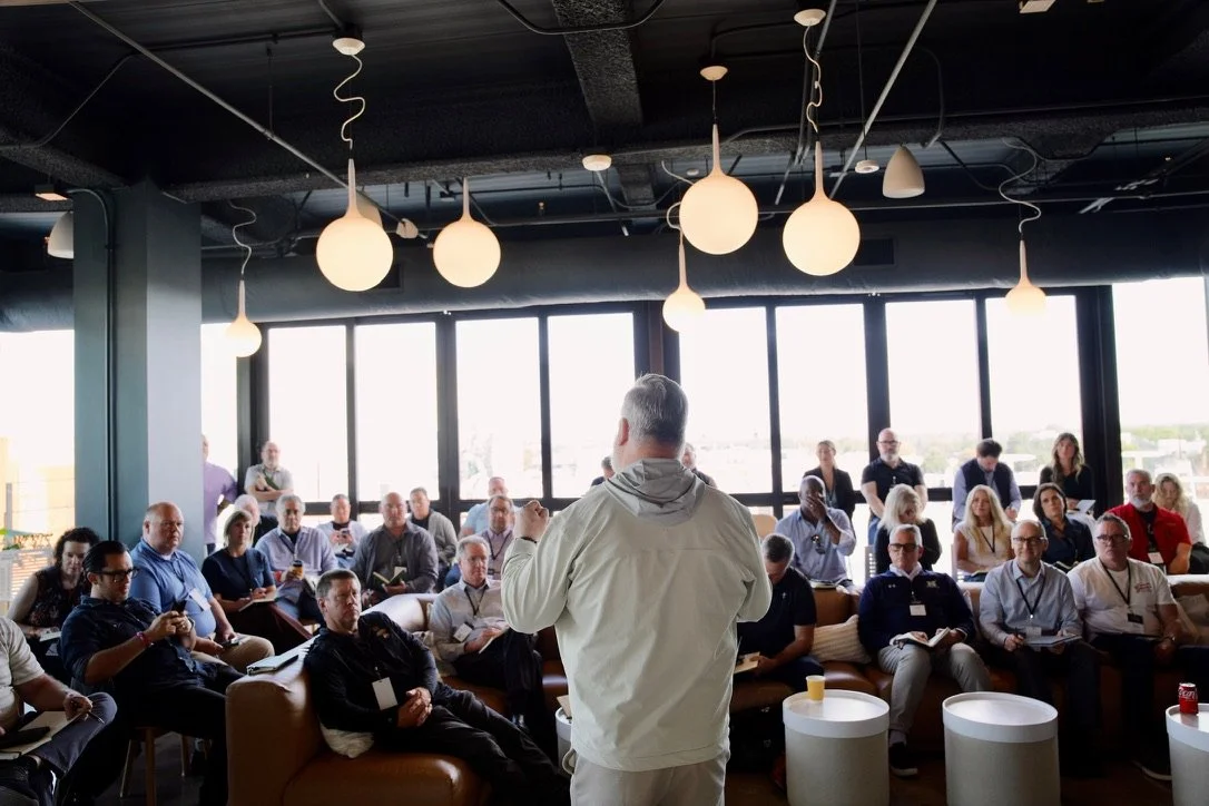 A man giving a presentation or speech in front of a seated audience in a modern, well-lit room with large windows and hanging round light fixtures.