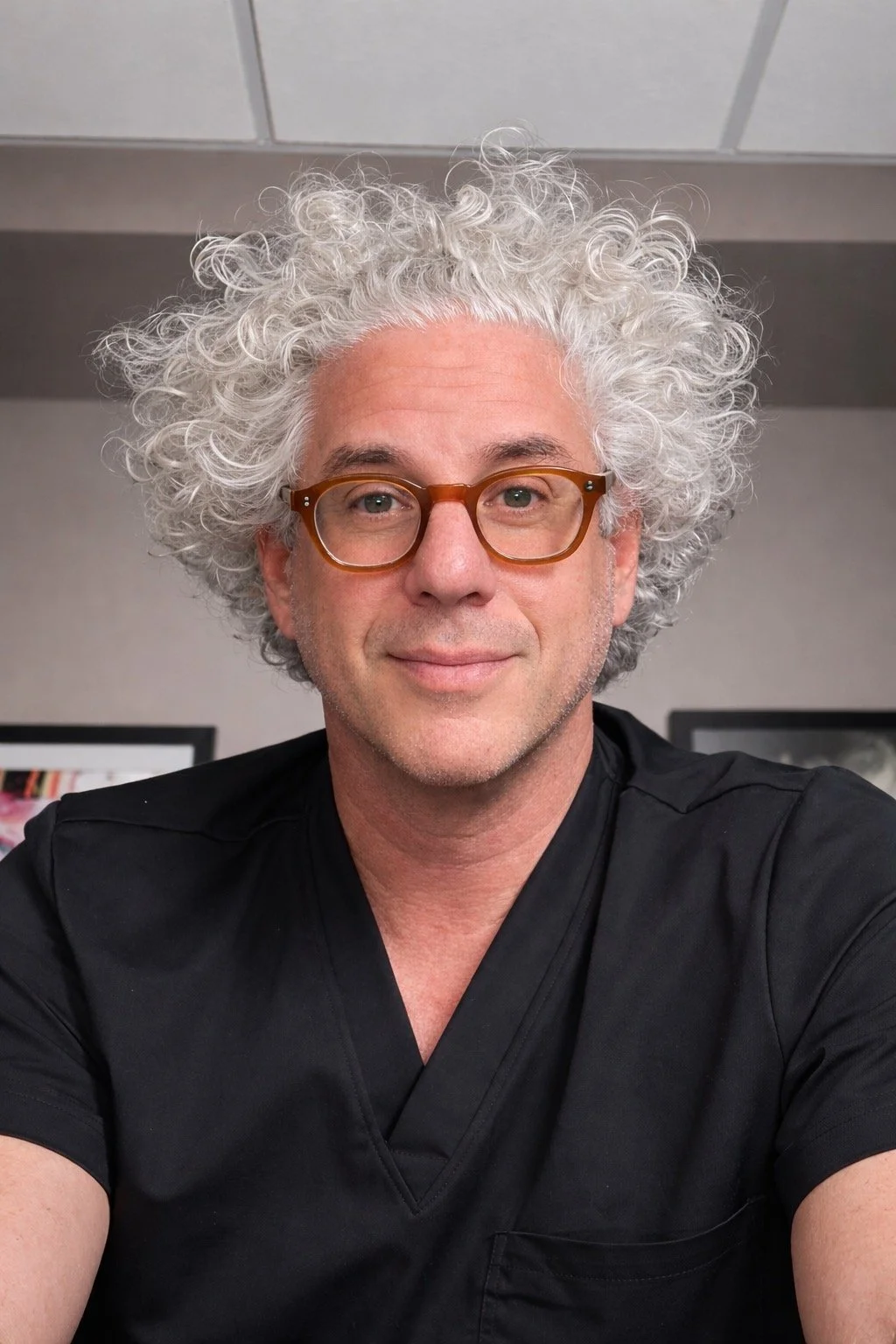 A man with curly white hair, glasses, and wearing a black shirt is smiling at the camera with a neutral background.
