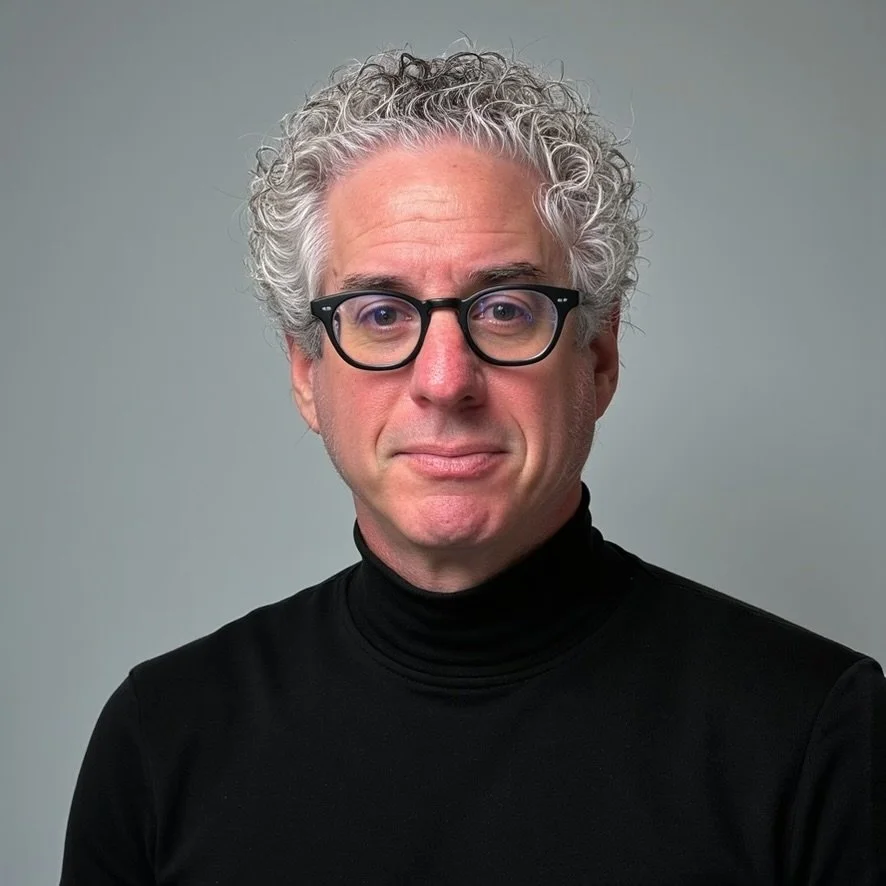 Portrait of a middle-aged man with curly gray hair, wearing black glasses and a black turtleneck, against a plain gray background.