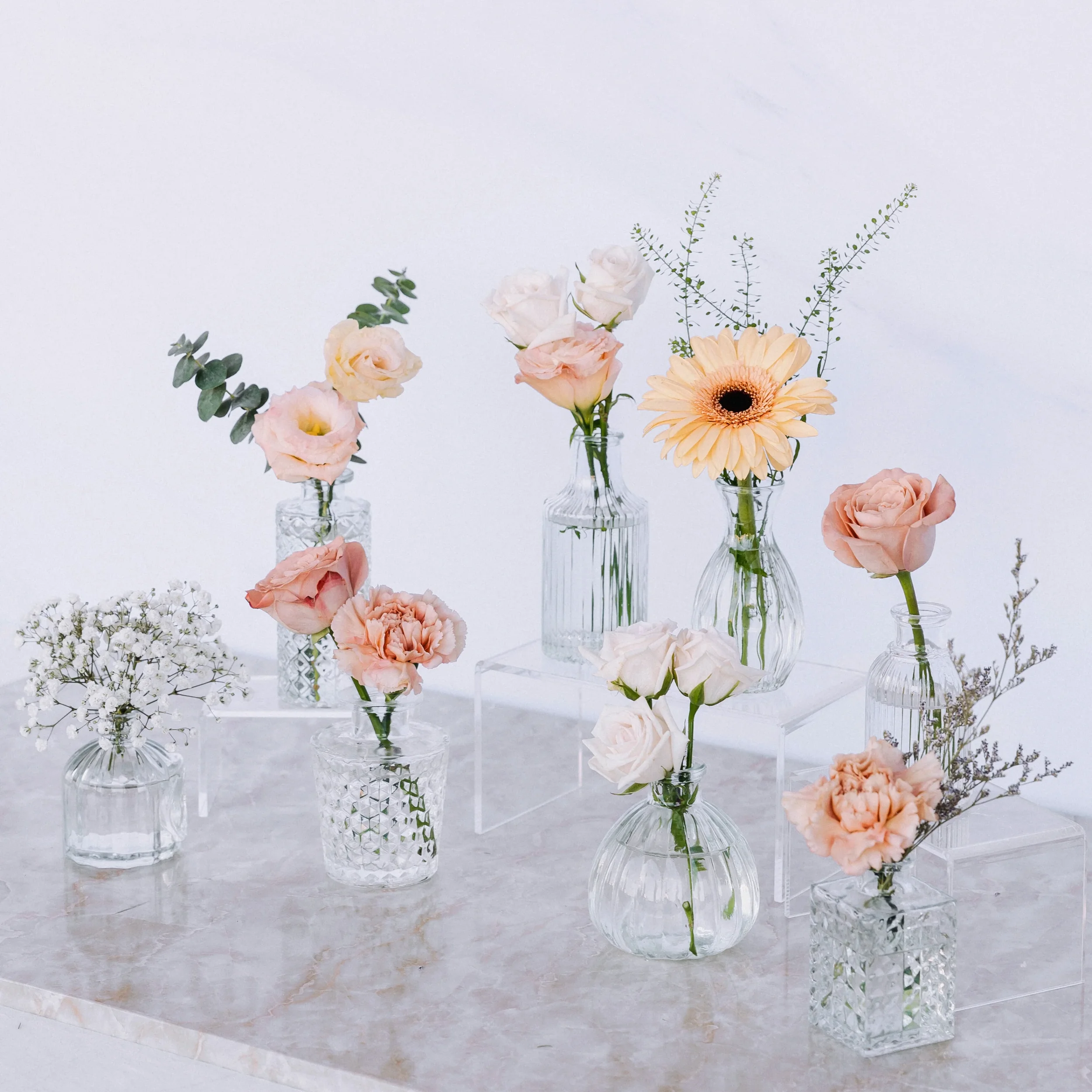 Various flowers in small glass vases arranged on a marble surface against a white background.