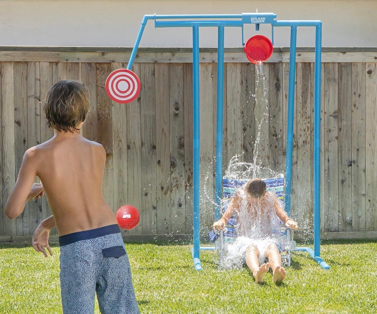 Two children playing outside in a backyard, with one on a water slide and the other standing nearby. The water slide has a large red bucket pouring water and a target prop in the background. Wooden fence surrounds the yard.