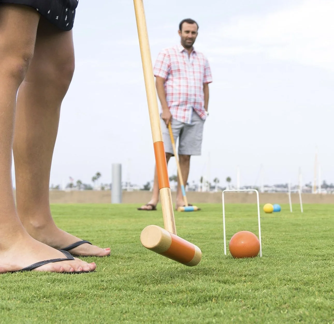 Close-up of people's feet and legs on a golf course playing mini golf, with a man in a plaid shirt and shorts holding a golf club in the background.