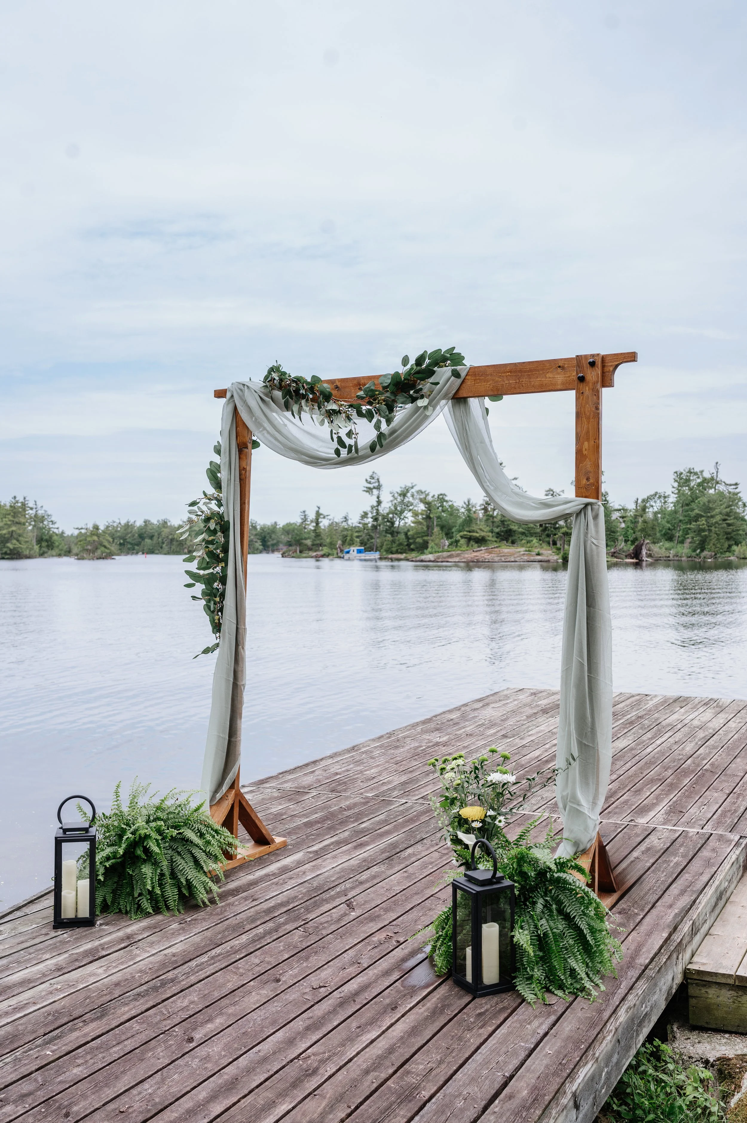 A wooden wedding arch decorated with greenery and white fabric, located on a dock by a lake, with lanterns and flowers at the base.